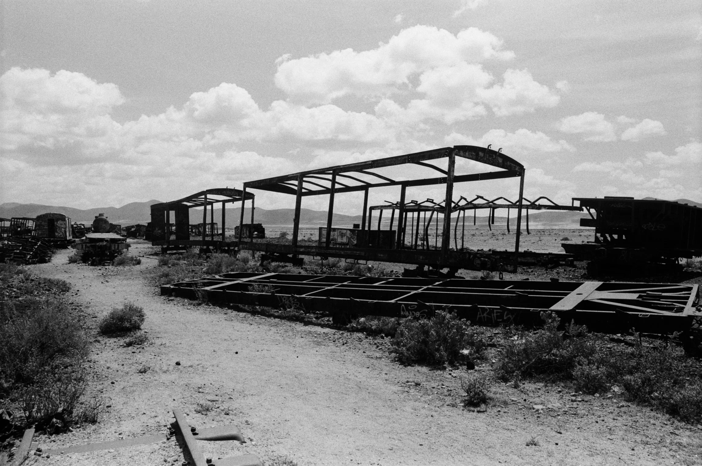Black and white photo of abandoned, dilapidated train cars in a desert landscape, with sparse vegetation and mountains in the background.
