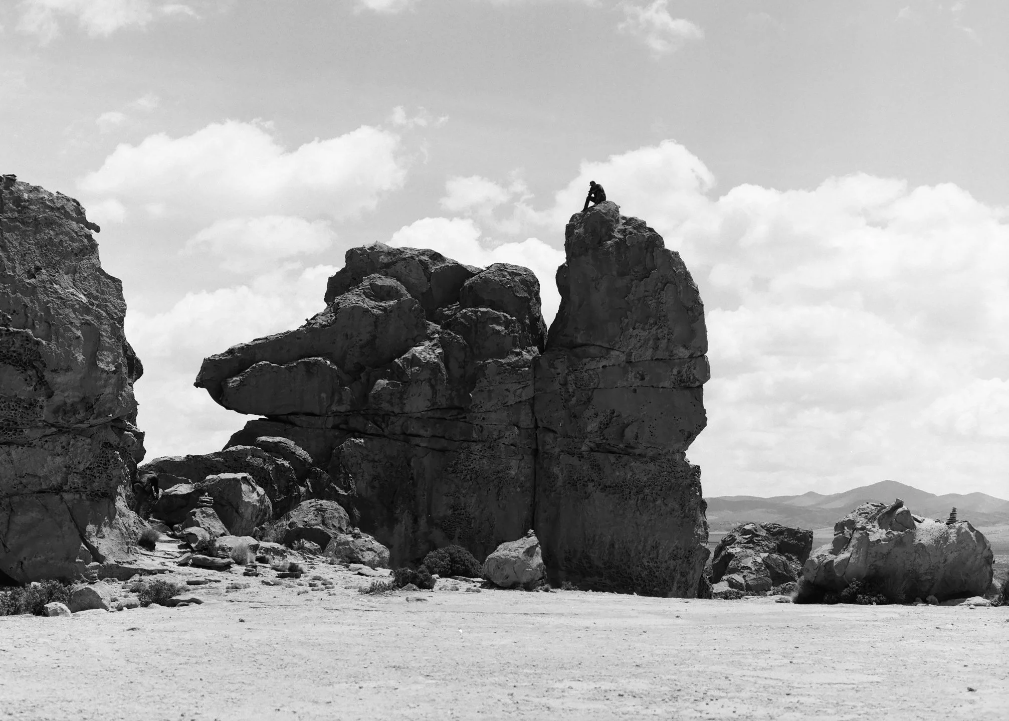 A person sits on top of a large rock formation in a desert landscape, with smaller rocks and boulders scattered around, and mountains in the distance under a sky filled with clouds.