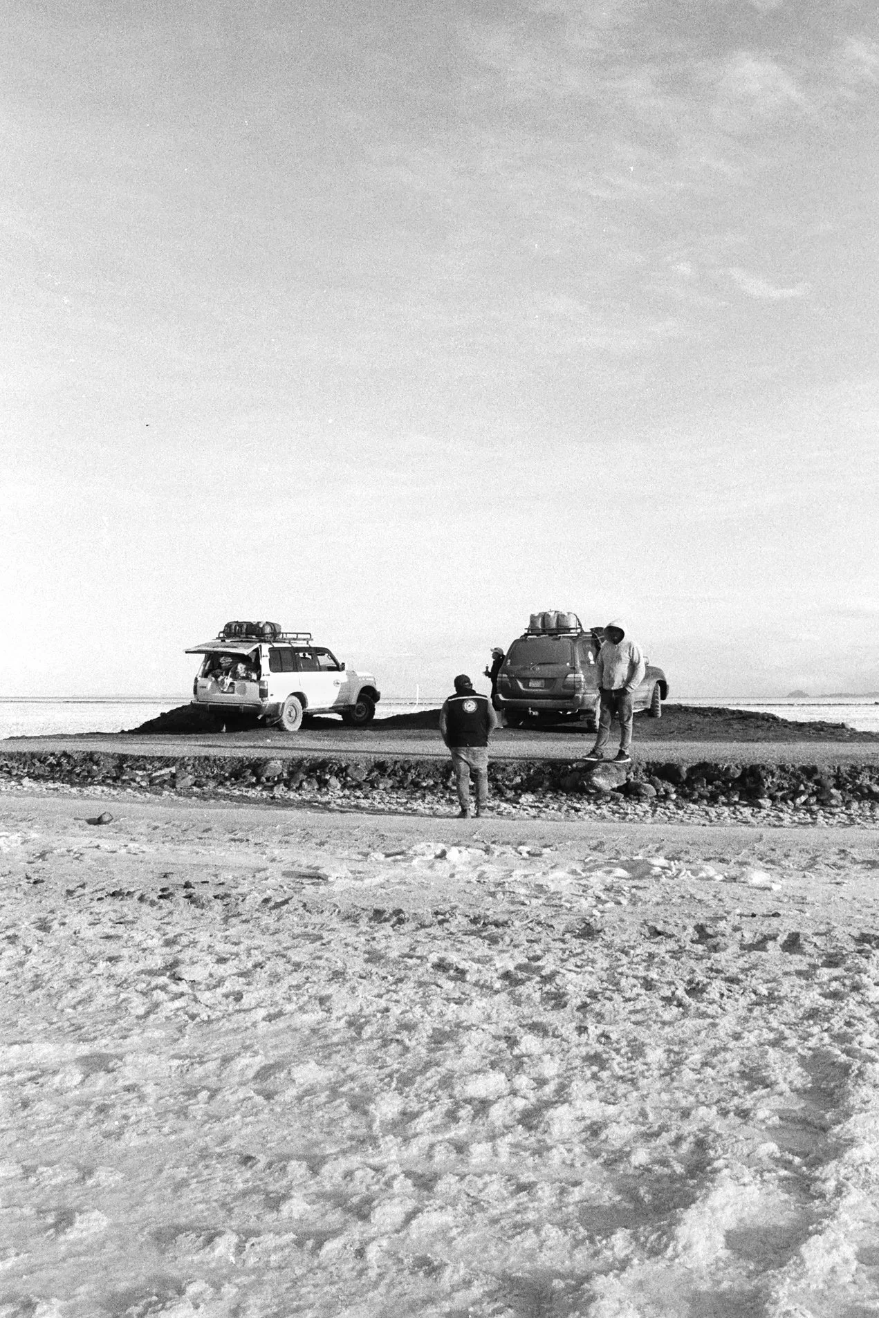Three people near two off-road vehicles on a dirt road in a flat, open landscape with a mostly clear sky.
