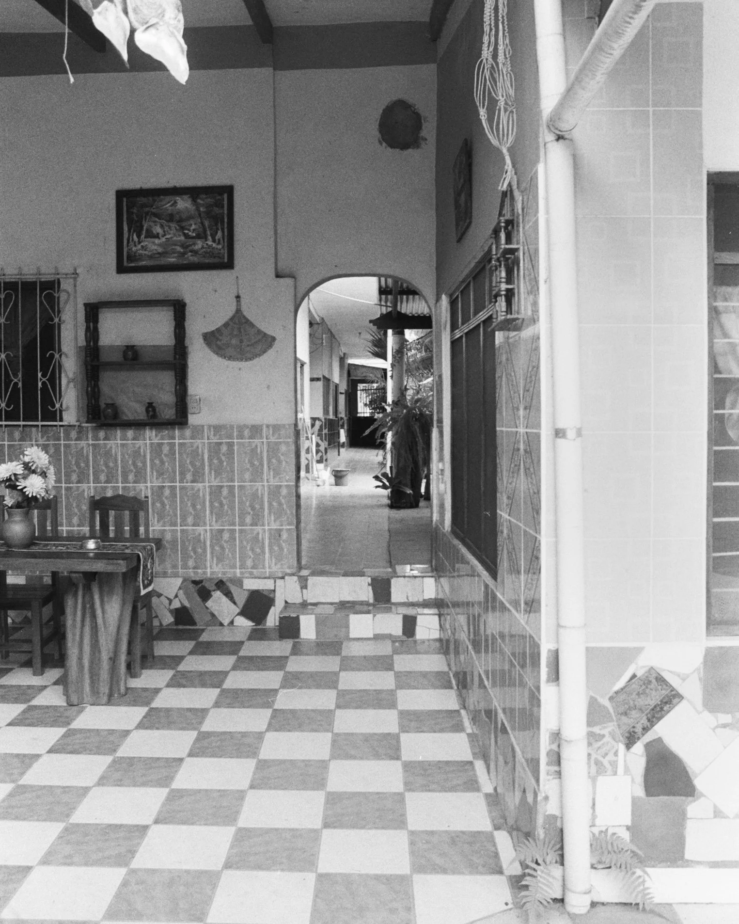 Interior view of a tiled room with a checkered floor, open doorway leading to an outdoor area with potted plants, wooden furniture with flowers, and decorative wall art.