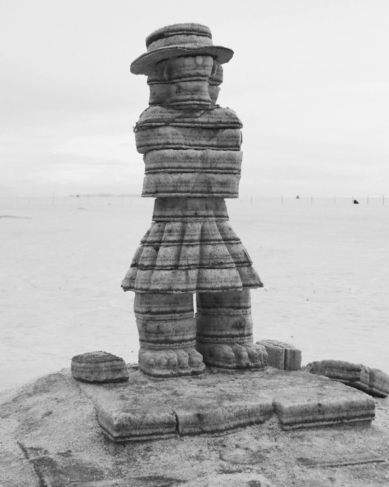 Black and white photo of a stack of stone sculptures resembling a person wearing a hat, standing on a stone platform by the water.