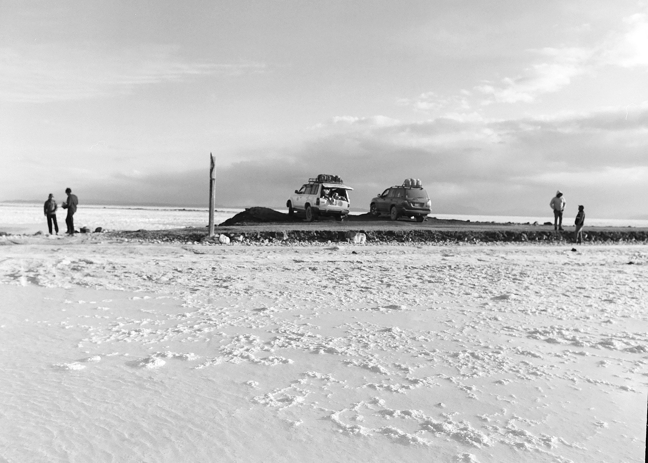 Black and white photo of a barren landscape with an elevated mound of dirt or gravel in the center, with two parked SUVs on top of it. Four people are visible, two on the left and two on the right, standing and talking near the vehicles. The sky is p