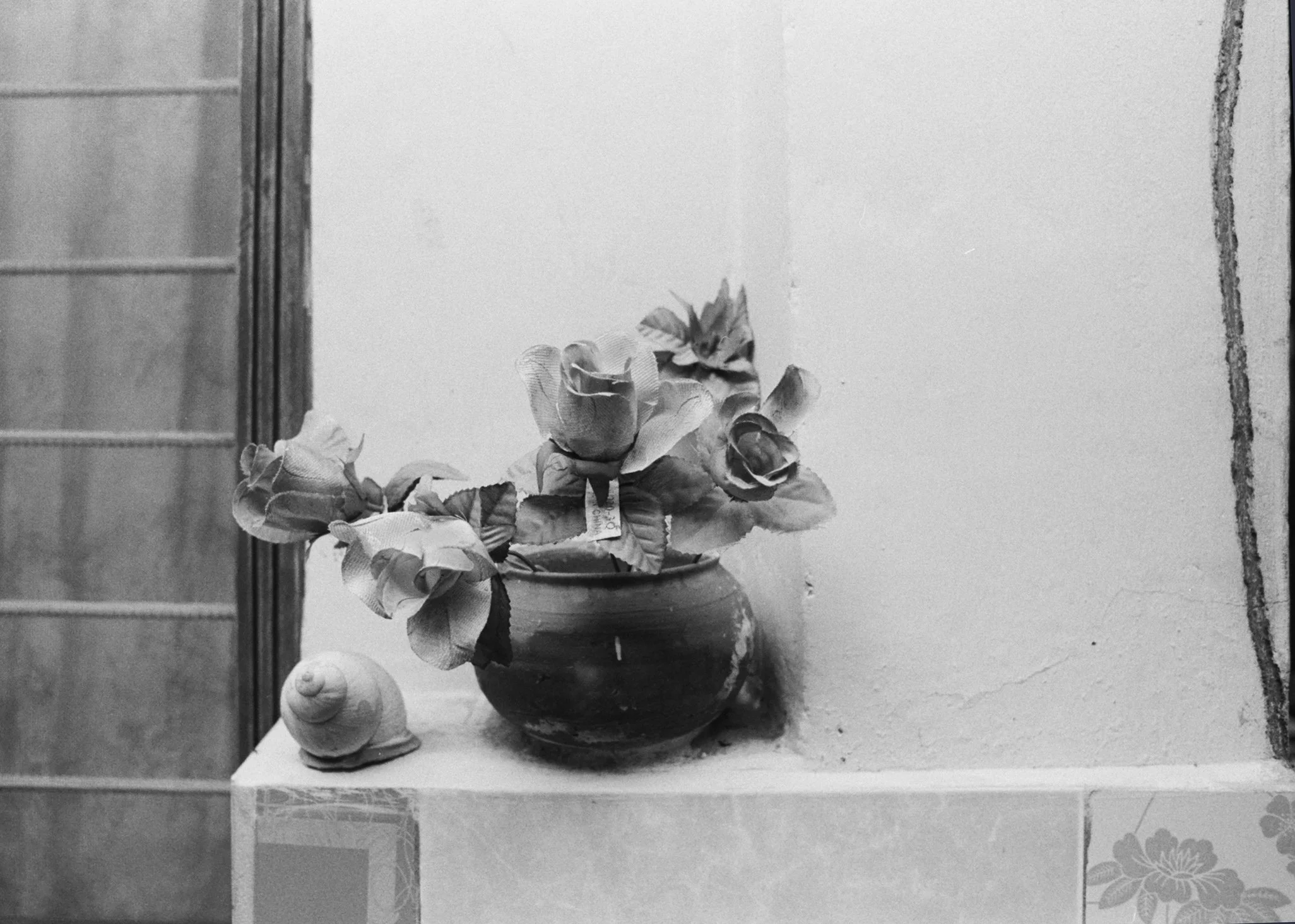 A still life black and white photo of a vase with artificial roses and a seashell on a ledge, against a plain wall with part of a tiled wall or door frame on the left.