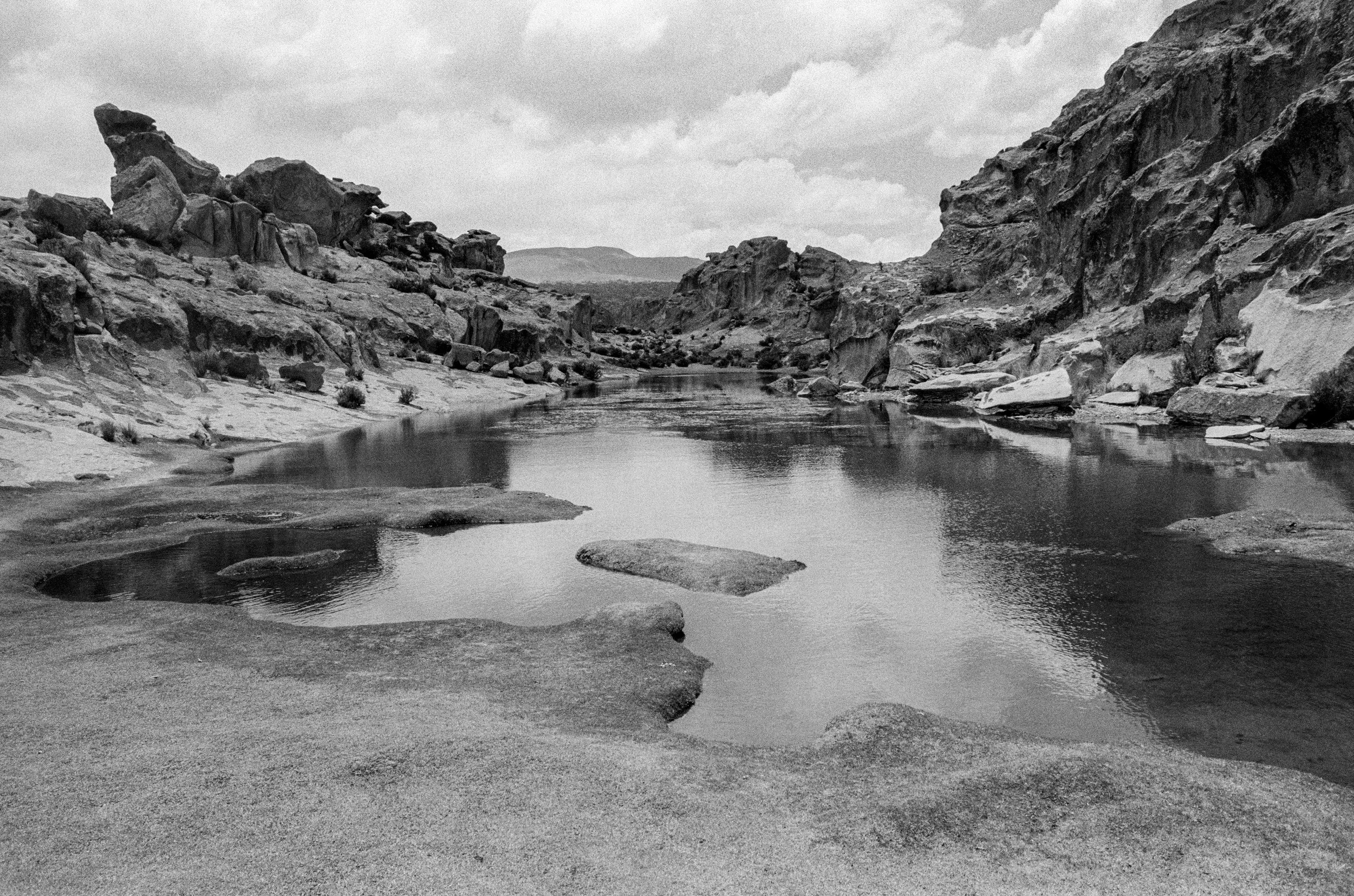 Black and white photo of a rocky river canyon with a calm river flowing through it and large cliff formations on both sides, under a partly cloudy sky.