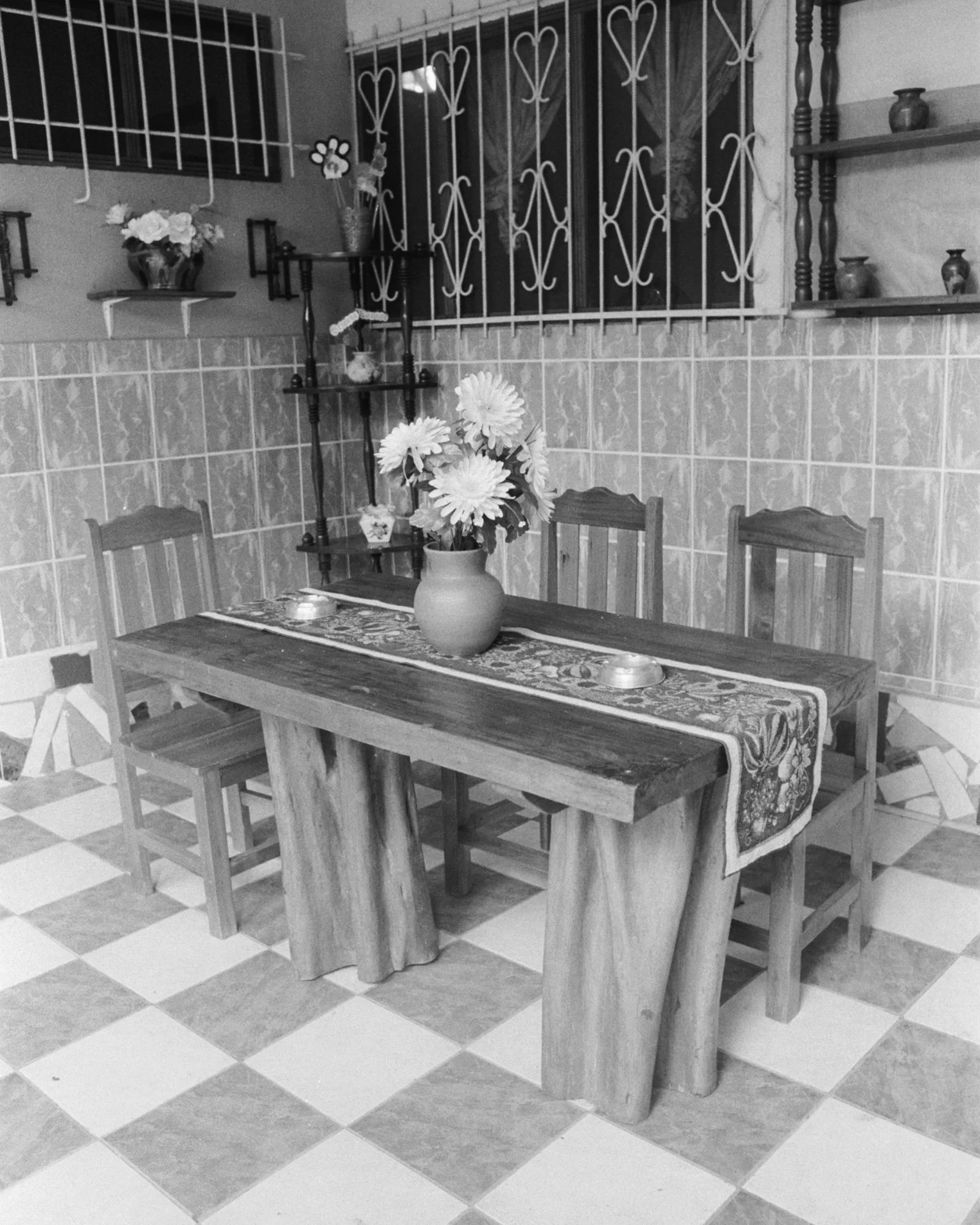 A wooden dining table with a floral table runner and a vase of large flowers, surrounded by four chairs, in a tiled room with decorative shelving and iron window bars.