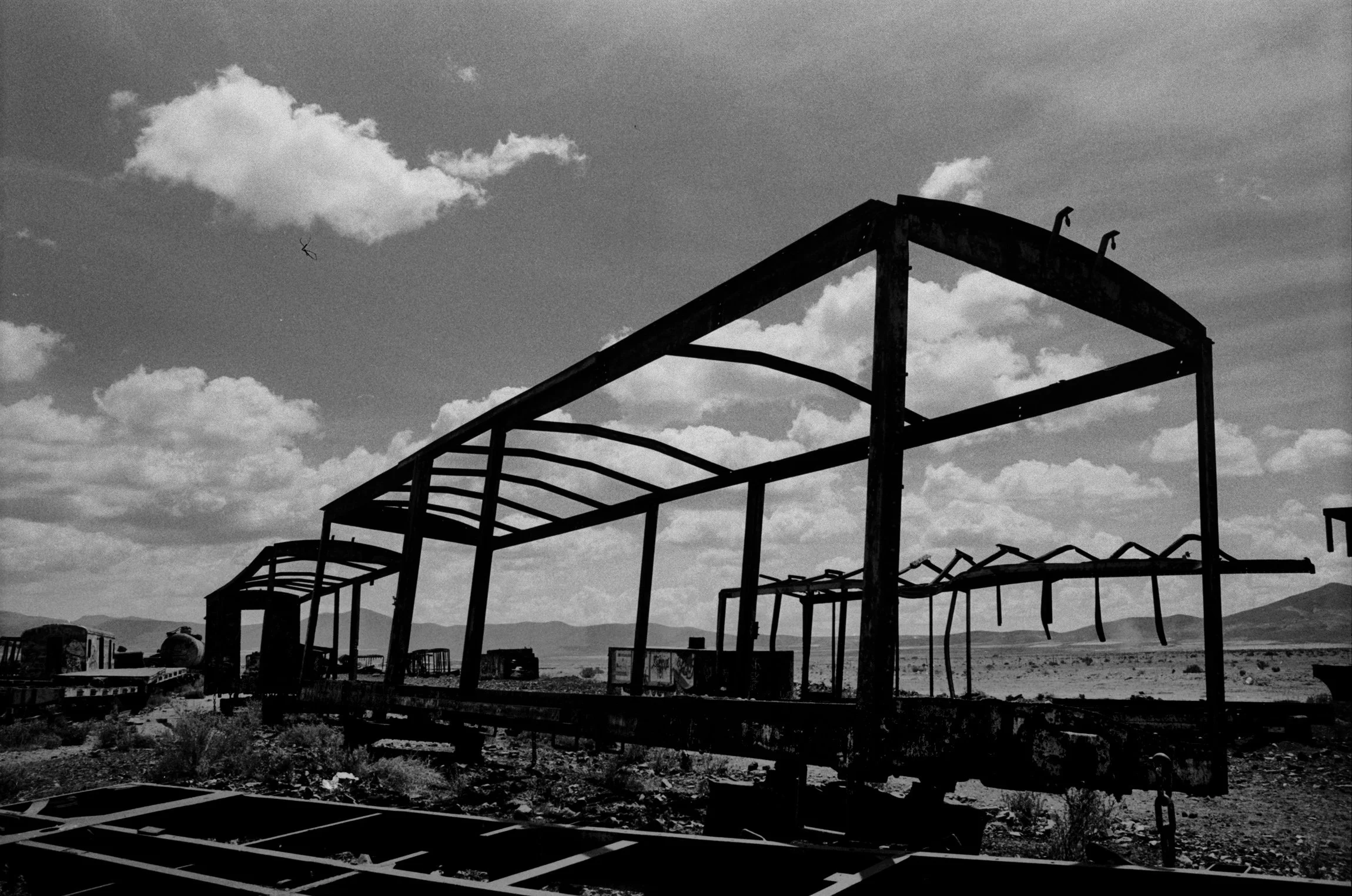 Black and white photo of a dilapidated, empty railroad car frame in a desert landscape with clouds in the sky.