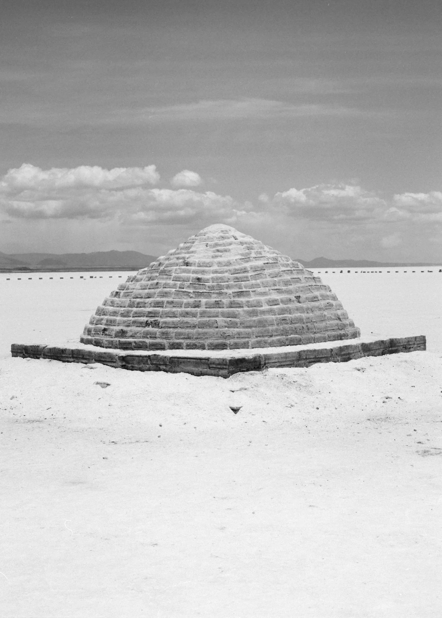 A brick and concrete circular structure in a desert landscape with a distant mountain range and cloudy sky.