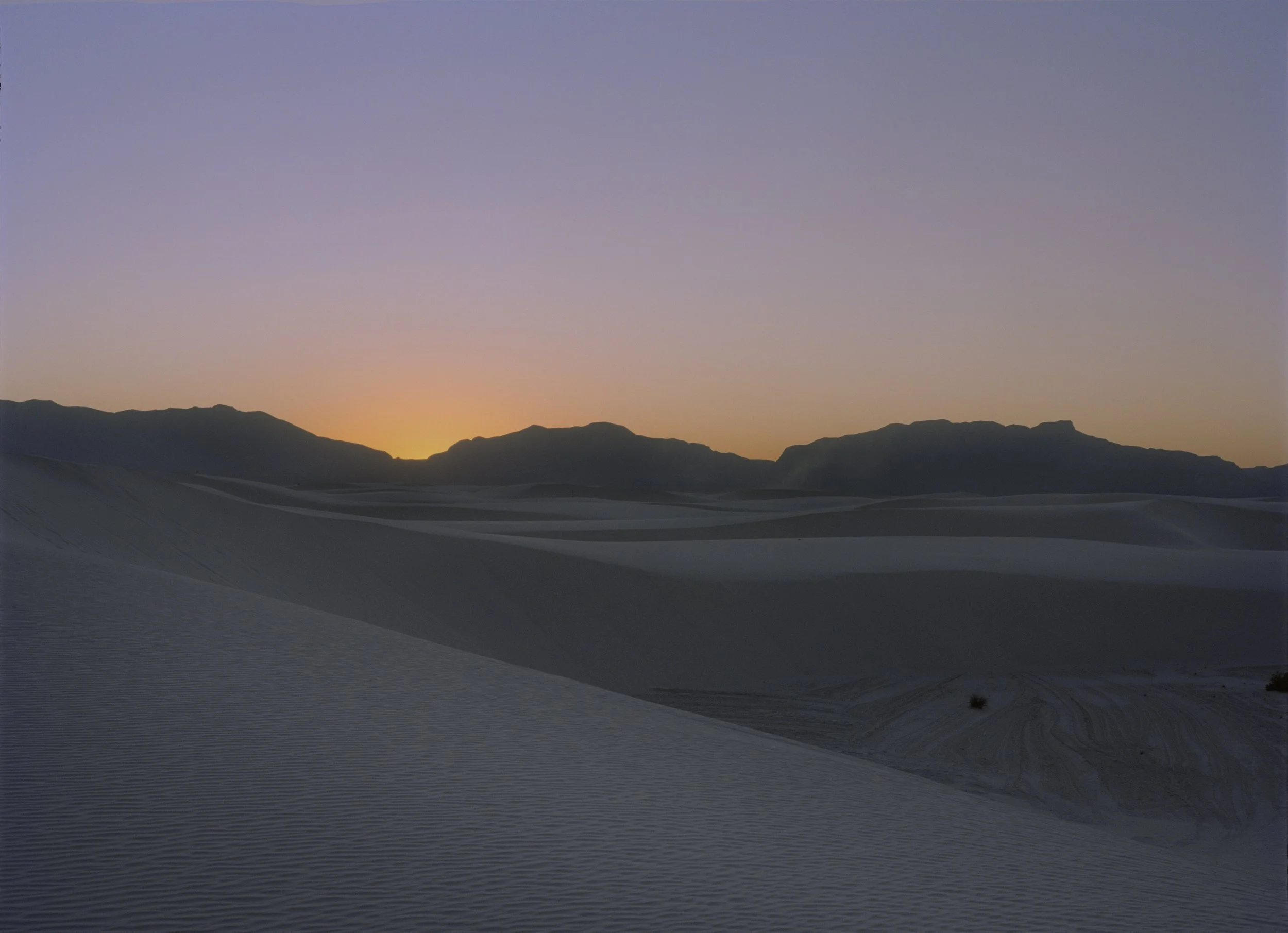 Desert landscape at sunset with sand dunes and mountains in the background, sky transitioning from orange to purple.