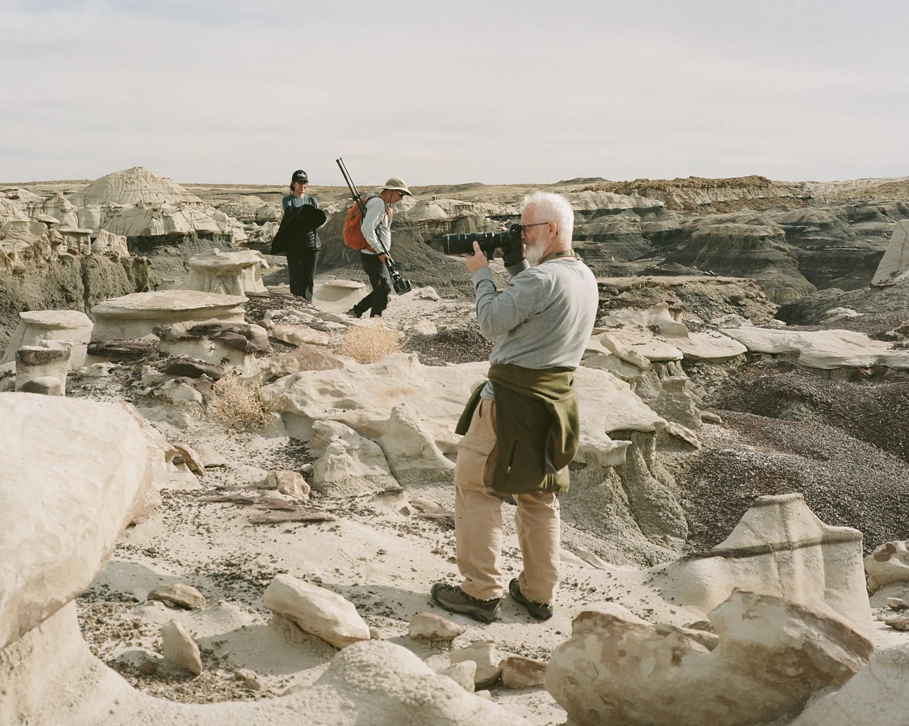 Three people exploring a rocky, desert landscape with unique formations, one older man with white hair and glasses taking photos, two other individuals carrying camera equipment and backpacks walking on uneven terrain.