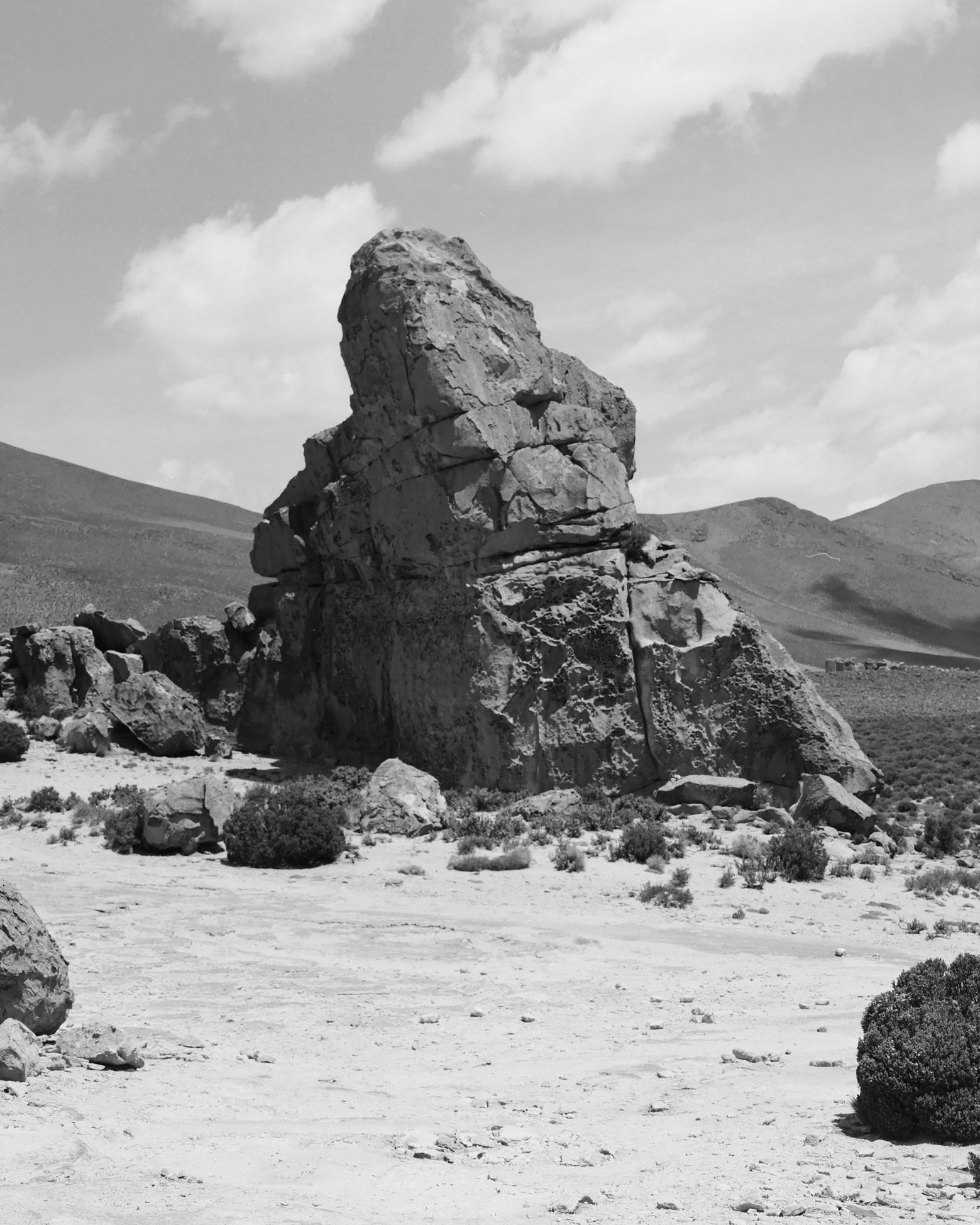 Black and white photograph of a large, uniquely shaped rock formation in a desert landscape with distant hills and a scattered cloud sky.