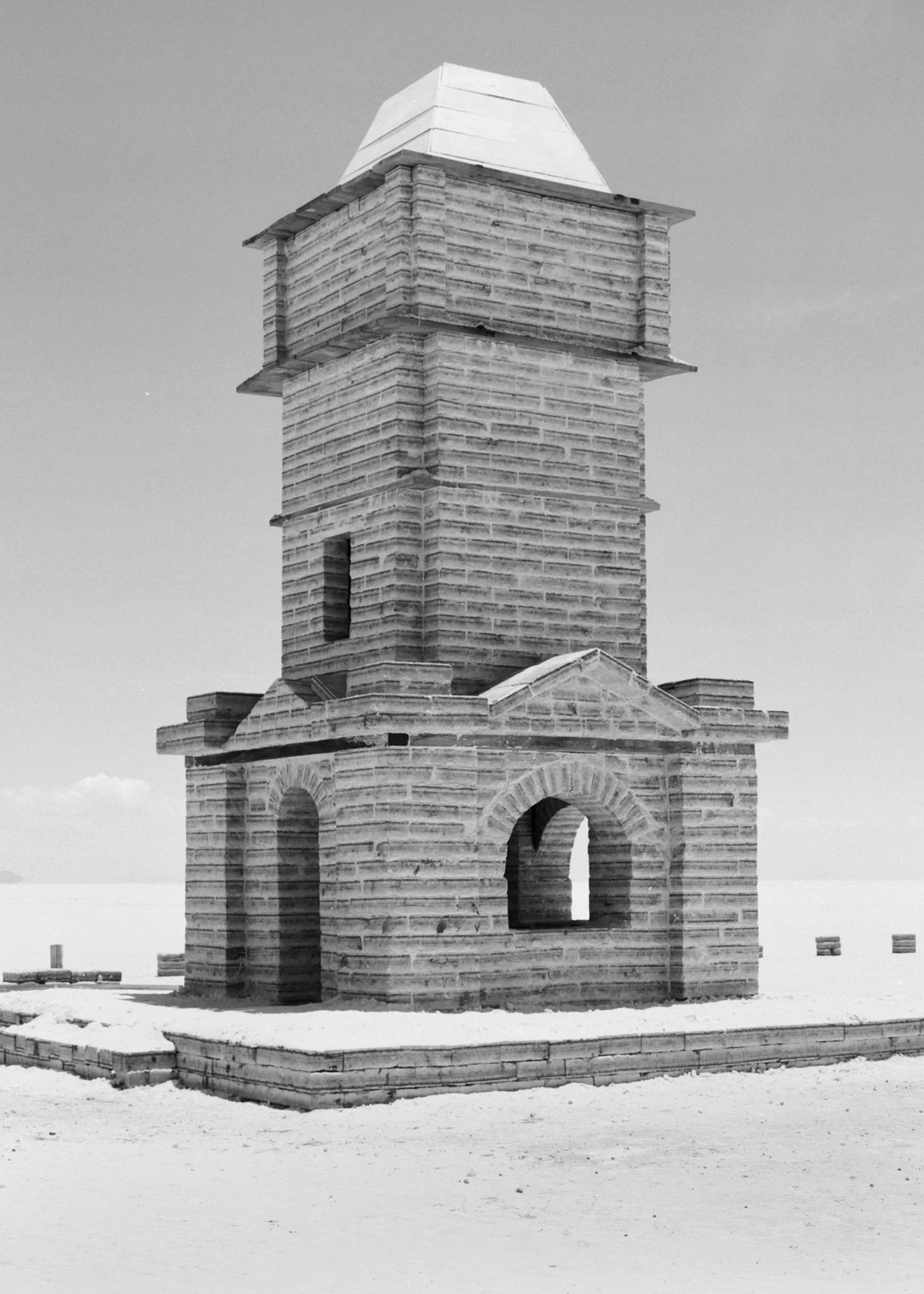A historic lighthouse structure made of brick and wood, with an archway at the base and a square tower on top, set in a snow-covered landscape.