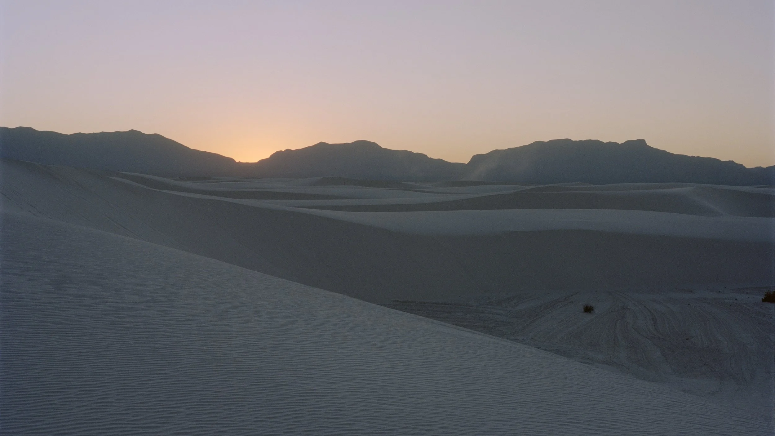 Sunset over sand dunes with mountains in the background under a clear sky.