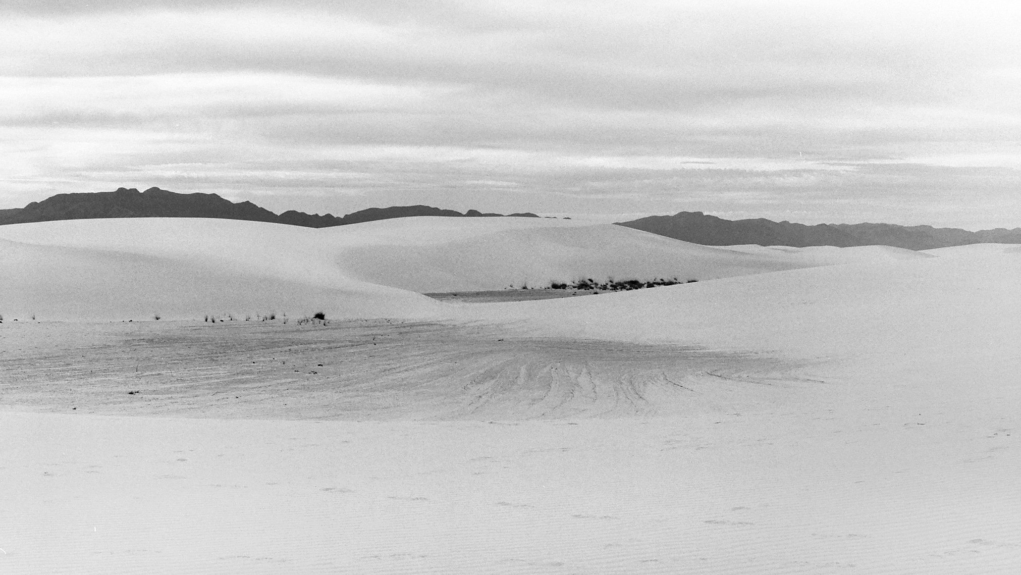 Black and white photo of sand dunes in a desert landscape with mountains in the background and a cloudy sky.