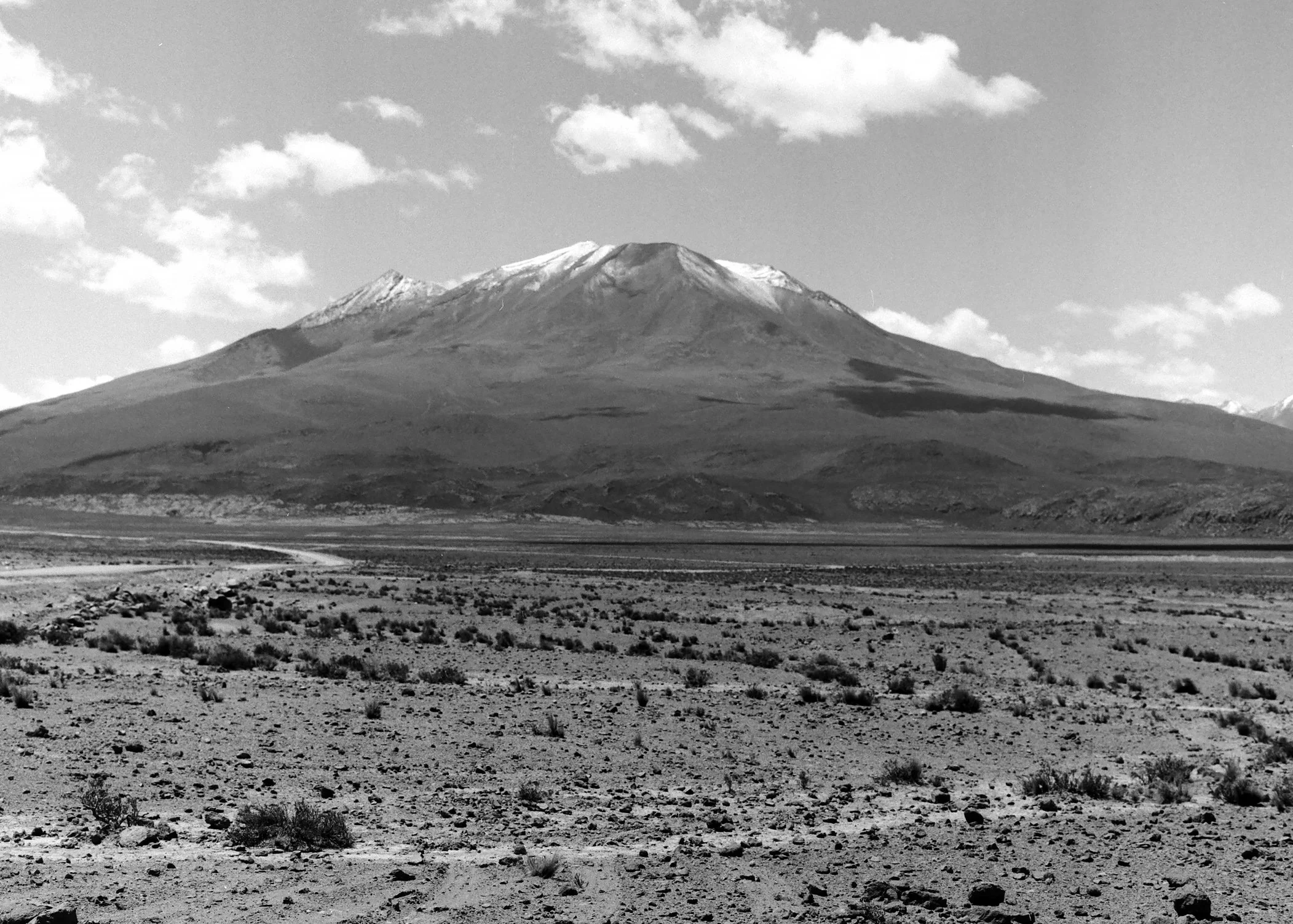 Black and white photo of a barren desert landscape with a tall mountain in the background, some clouds in the sky.