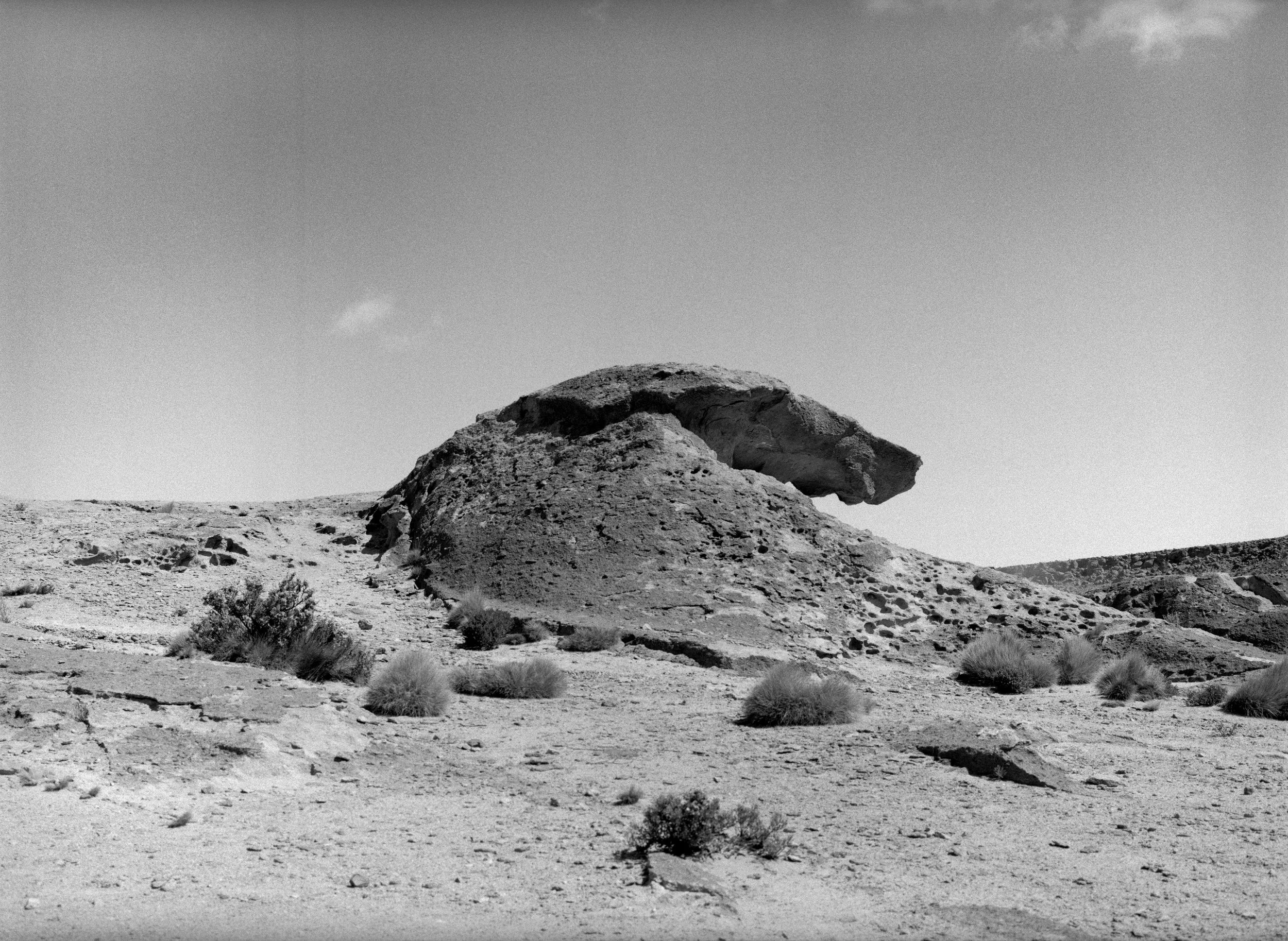A black and white photo of a desert landscape with a large, rounded rock formation in the center. Sparse desert vegetation is scattered across the foreground, and the sky is mostly clear with a few clouds.