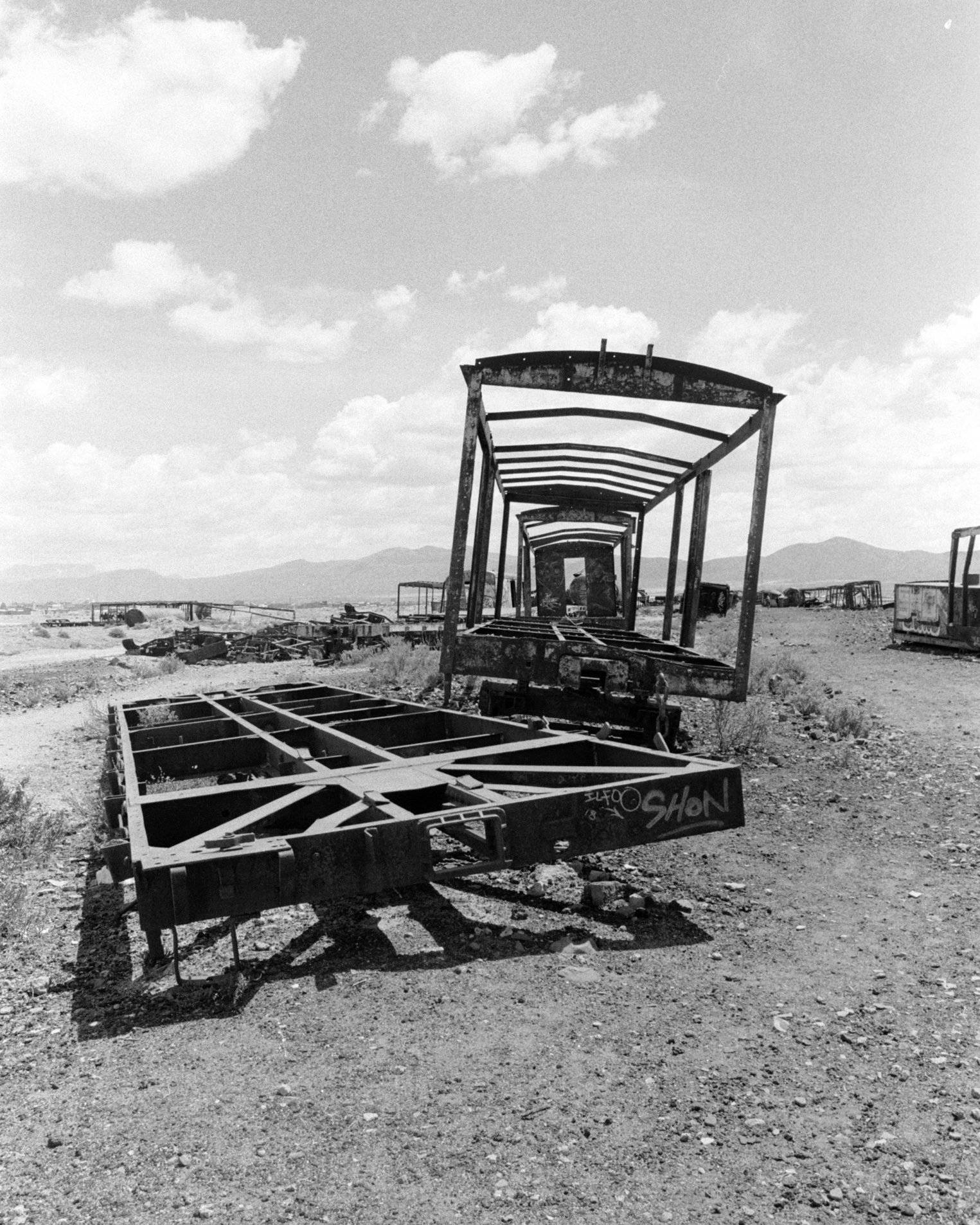 Black and white photo of abandoned, rusted, and broken-down railroad cars in a desert landscape with mountains in the background.