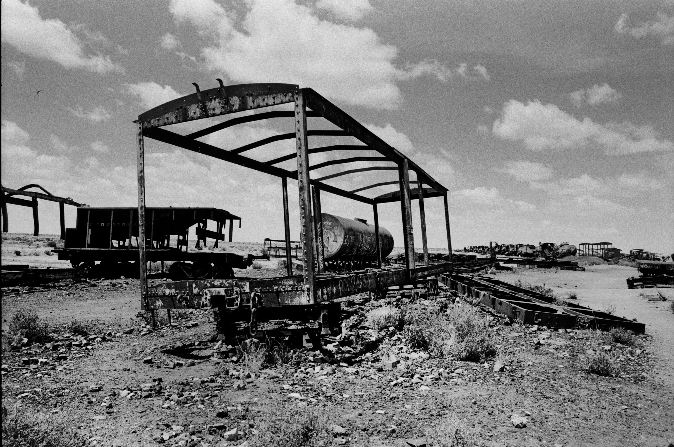 A black-and-white photo of abandoned, rusted train cars and structures in a desert landscape under a partly cloudy sky.