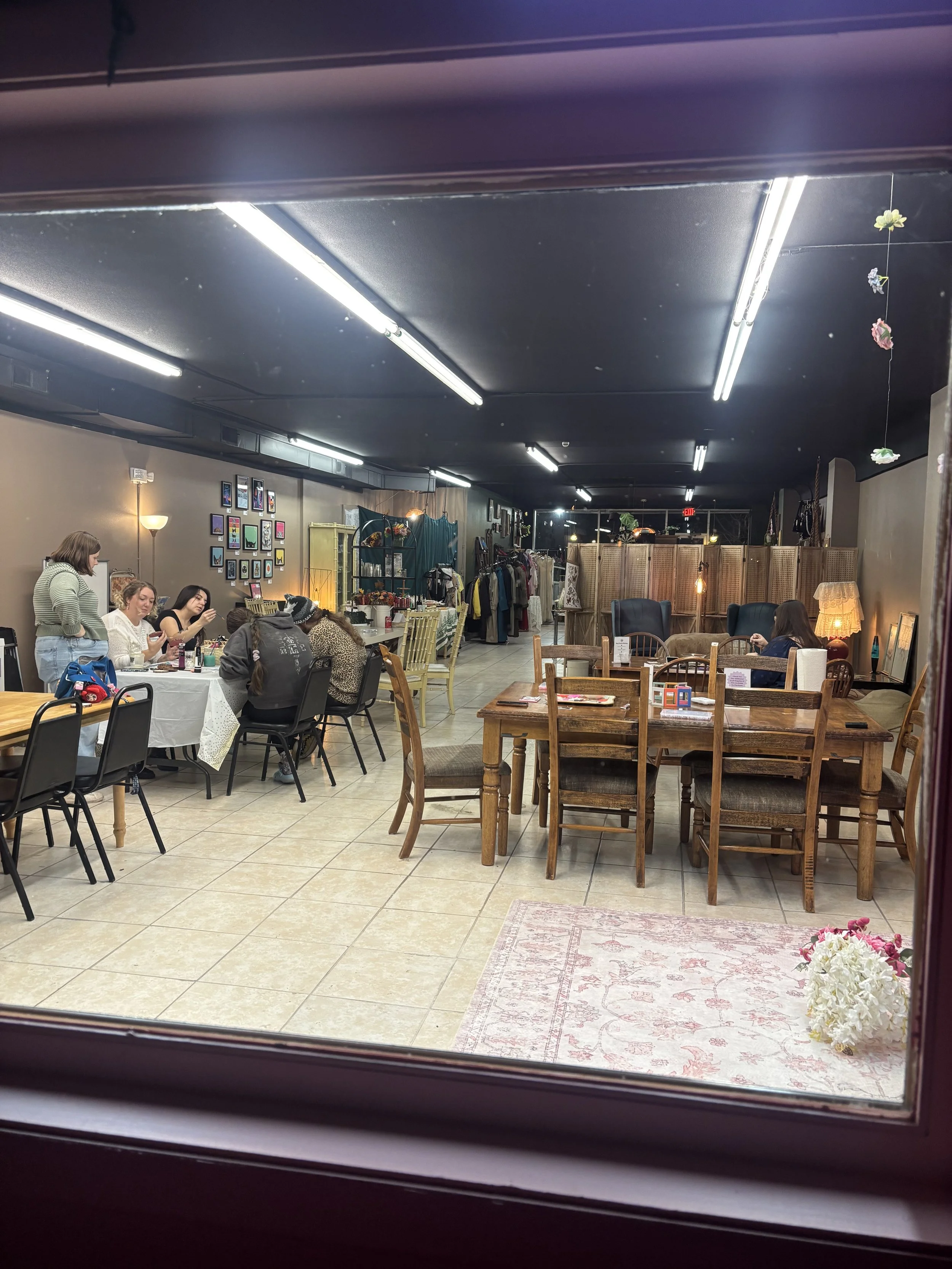 View of an indoor space seen through a window, with tables and chairs, some people sitting and some standing, a clothing rack, wall decorations, and soft lighting.