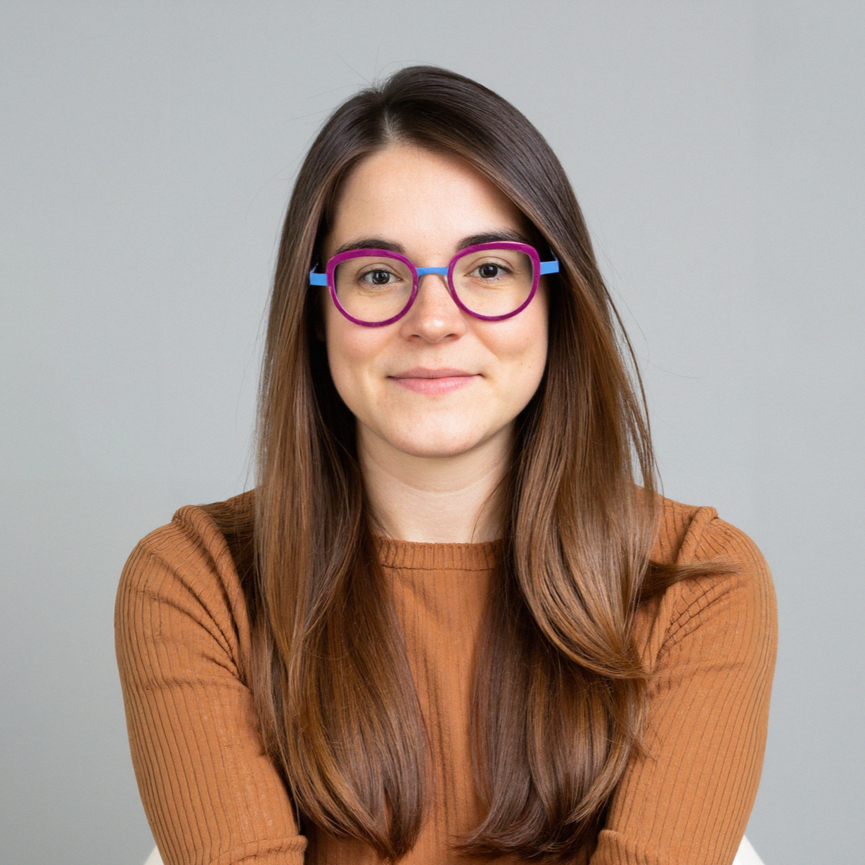 A woman with long brown hair wearing purple and blue eyeglasses and a brown sweater, smiling at the camera against a plain gray background.