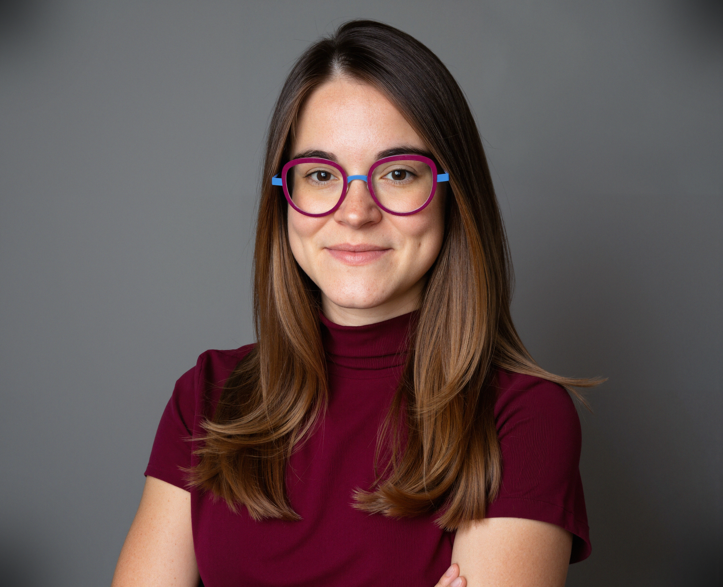Portrait of a woman with long brown hair wearing purple and blue glasses and a maroon top, smiling against a gray background.