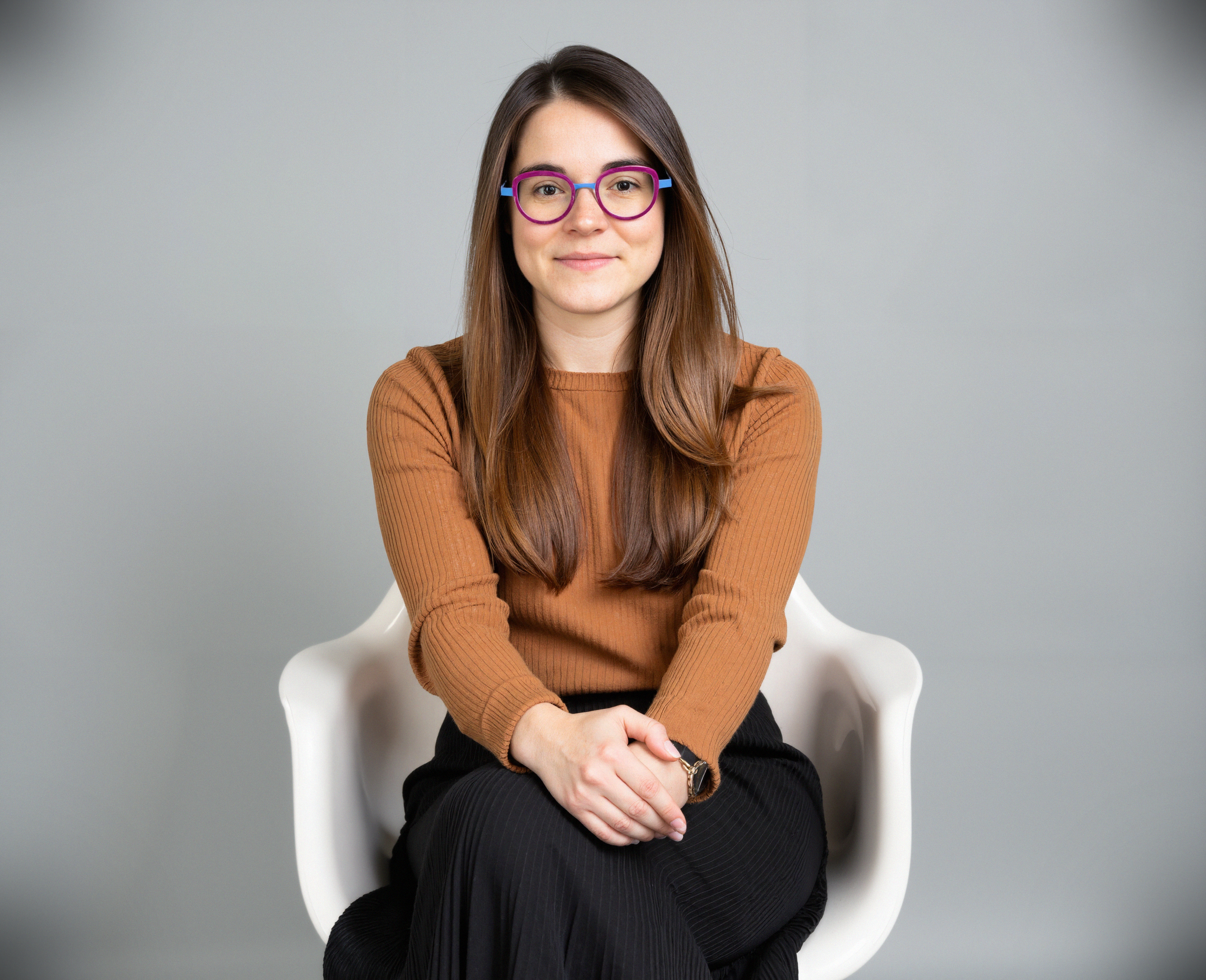 A woman with long brown hair wearing purple glasses, a brown long-sleeve shirt, and black skirt, sitting on a white chair with her hands folded on her lap, against a plain gray background.