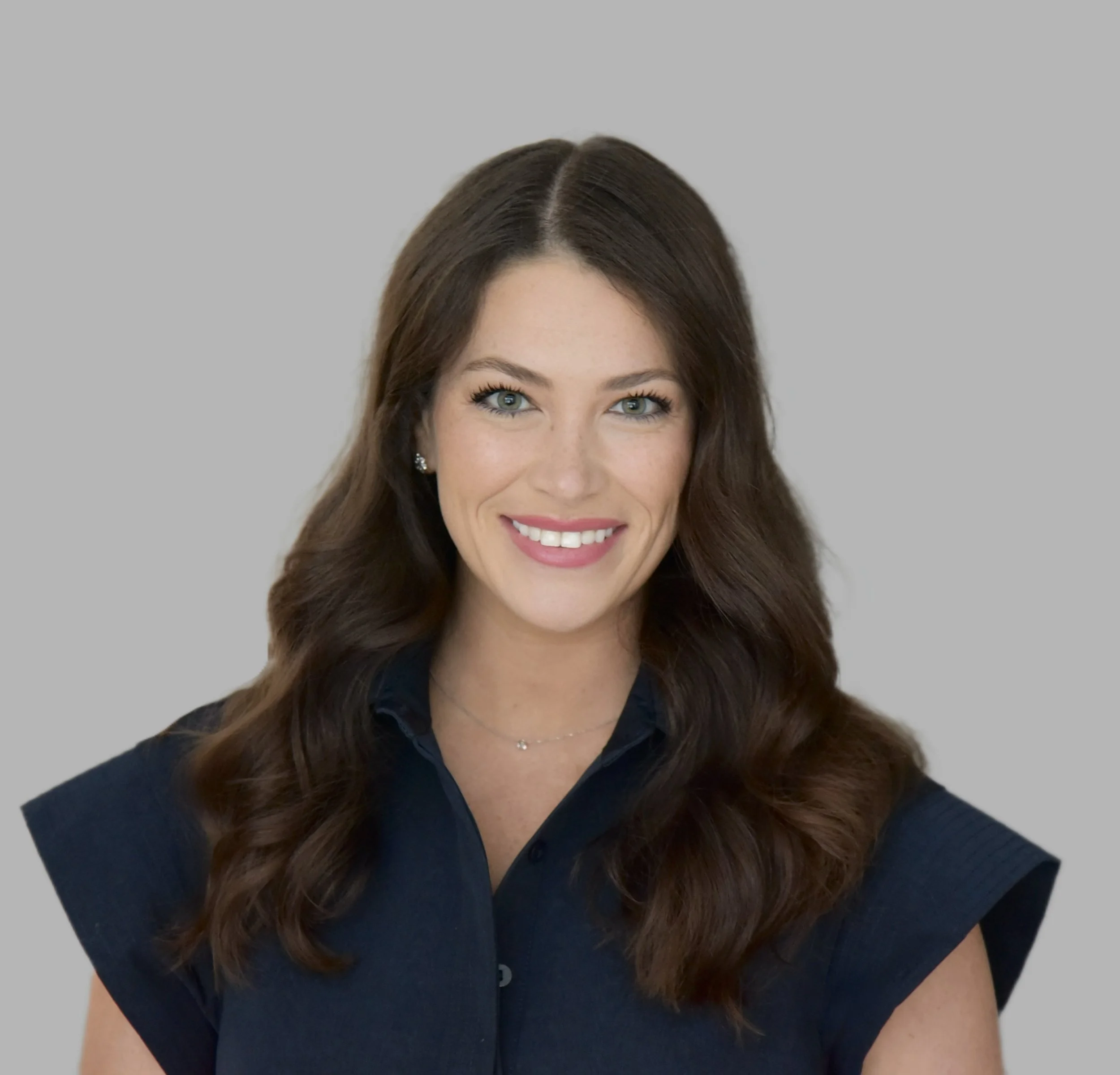 Close-up of a woman with long, wavy brown hair, blue eyes, and a bright smile, wearing a black top and small earrings, against a light gray background.