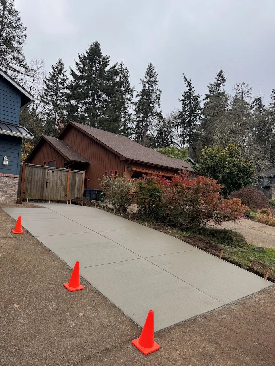 Freshly poured concrete drive with orange cones, in front of a red house with a sloped roof and surrounded by trees and shrubs.