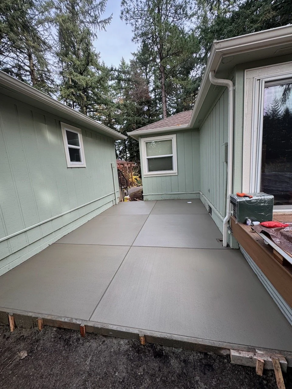 Newly poured concrete patio in the backyard of a house, surrounded by green siding and trees in the background.
