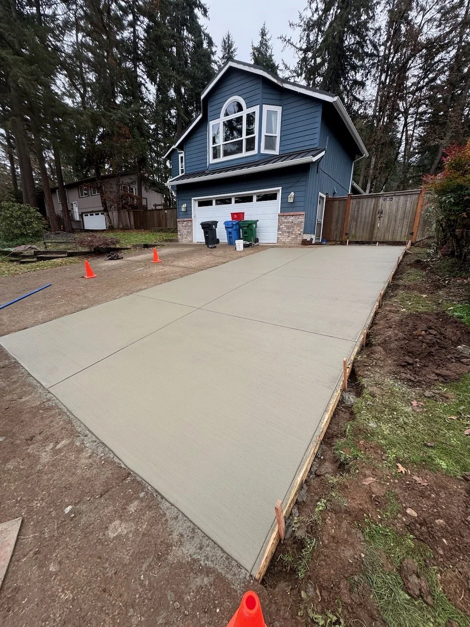 Freshly poured concrete driveway in front of a blue two-story house with a garage, surrounded by trees and construction cones.