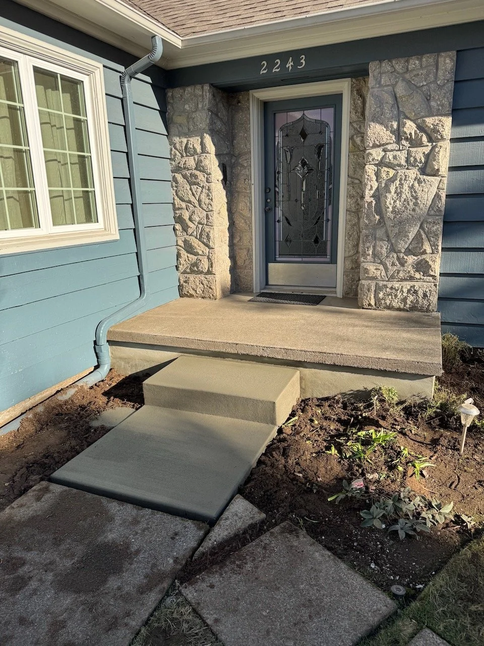 Front porch of a house with a glass door, stone accents, and house number 2243. There is a concrete ramp and steps leading to the door, with a small garden bed nearby.