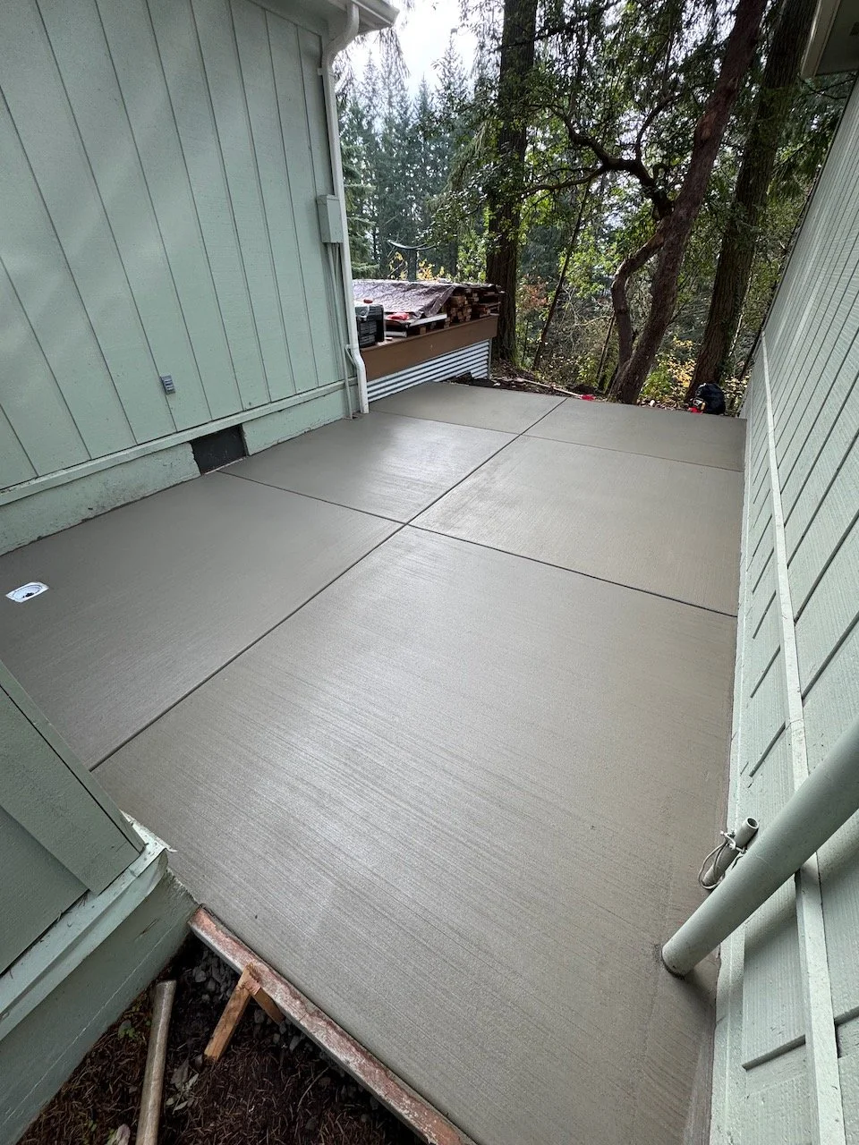 Newly poured concrete on a backyard porch with a view of trees and a woodpile in the background.