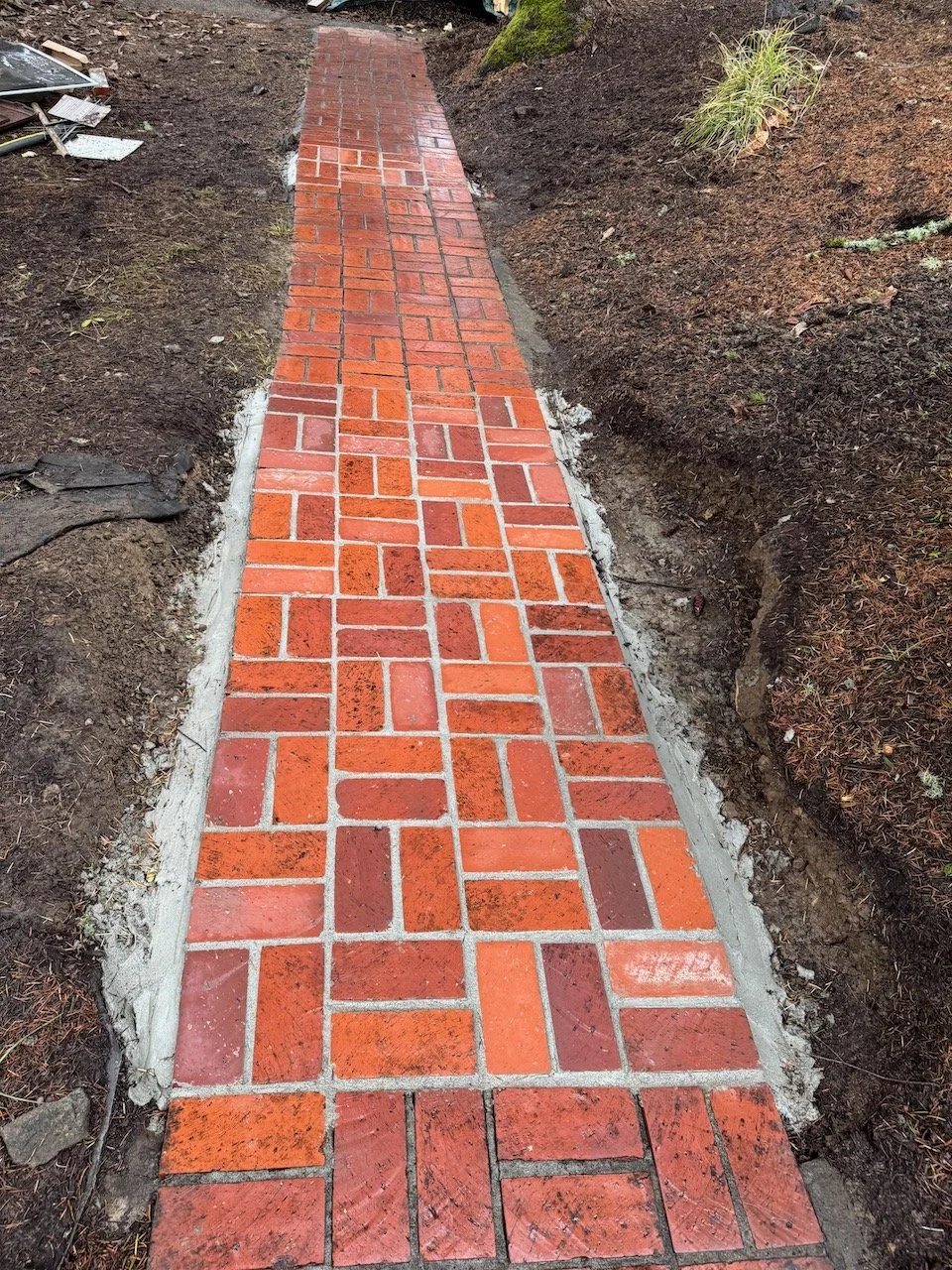 A newly paved brick sidewalk under construction, surrounded by dark soil and some plants.