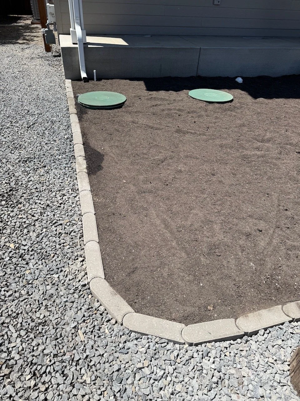 A small, rectangular dirt patch bordered by gray pavers, with two green circular covers near a house wall, and a gravel area to the left.