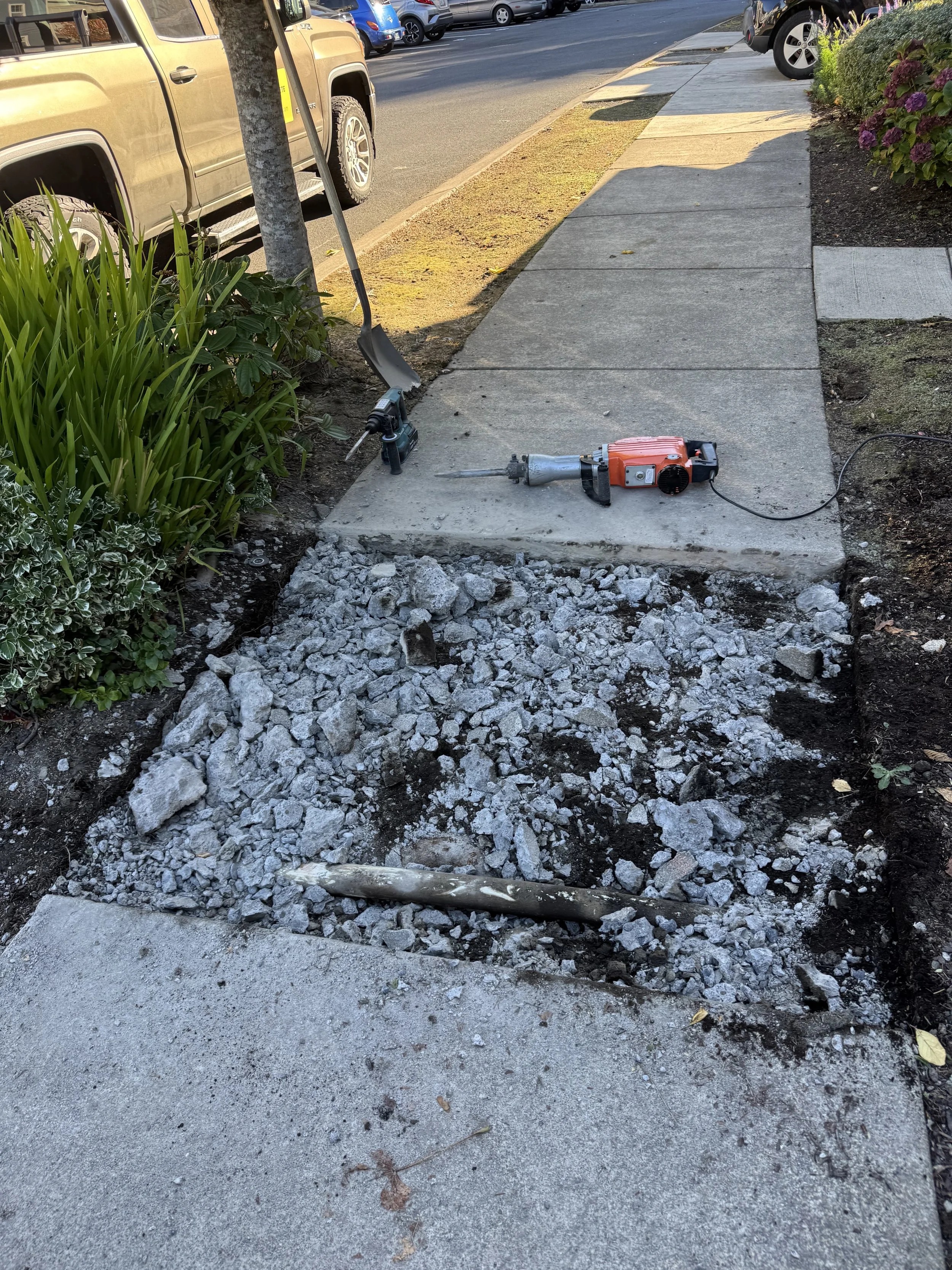 Construction work on a sidewalk shows a section of the sidewalk dug up with debris and tools, including a power sander, placed nearby.