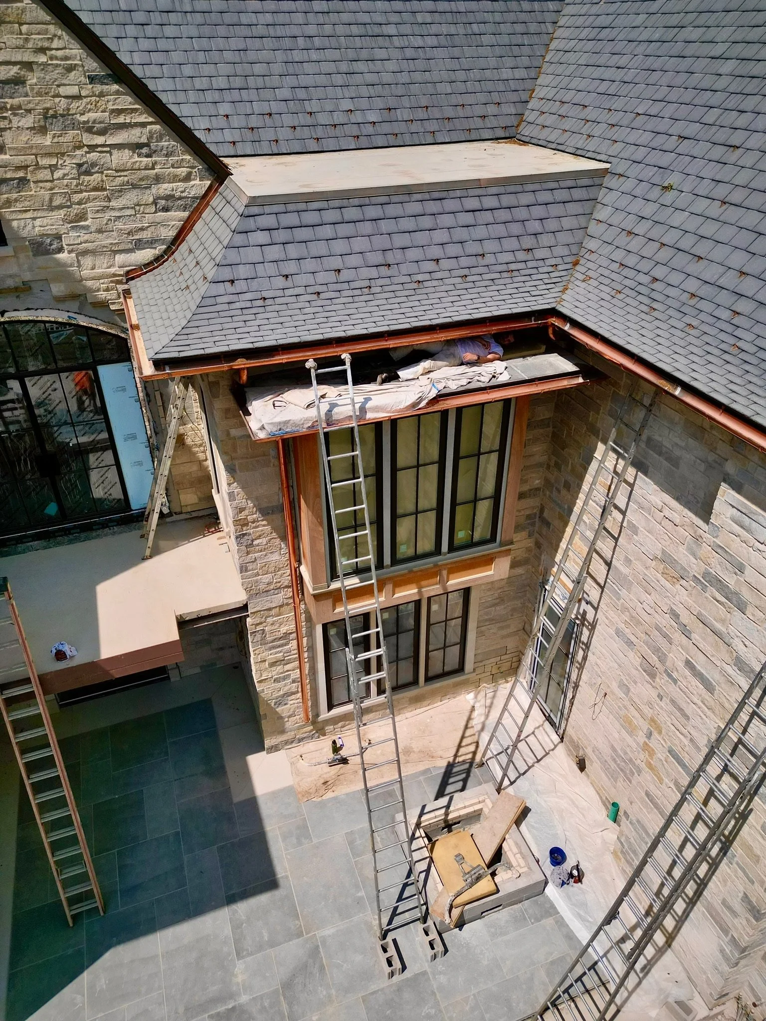 Construction workers installing a roof on a house's upper balcony, with ladders leaning against the building and tools scattered below.
