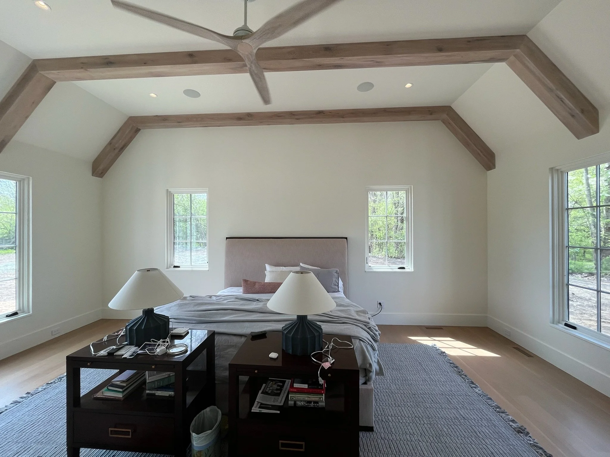 Unmade bed with nightstands and lamps in a bright bedroom with large windows and exposed wooden beams on the ceiling.