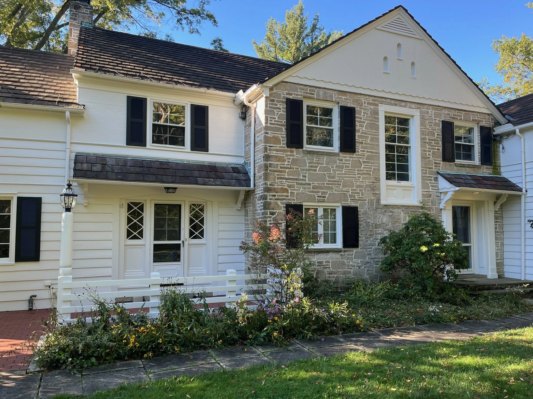 A two-story house with a combination of white siding and stone exterior, several windows with black shutters, a brick and shingle roof, and a small garden with flowers in front.