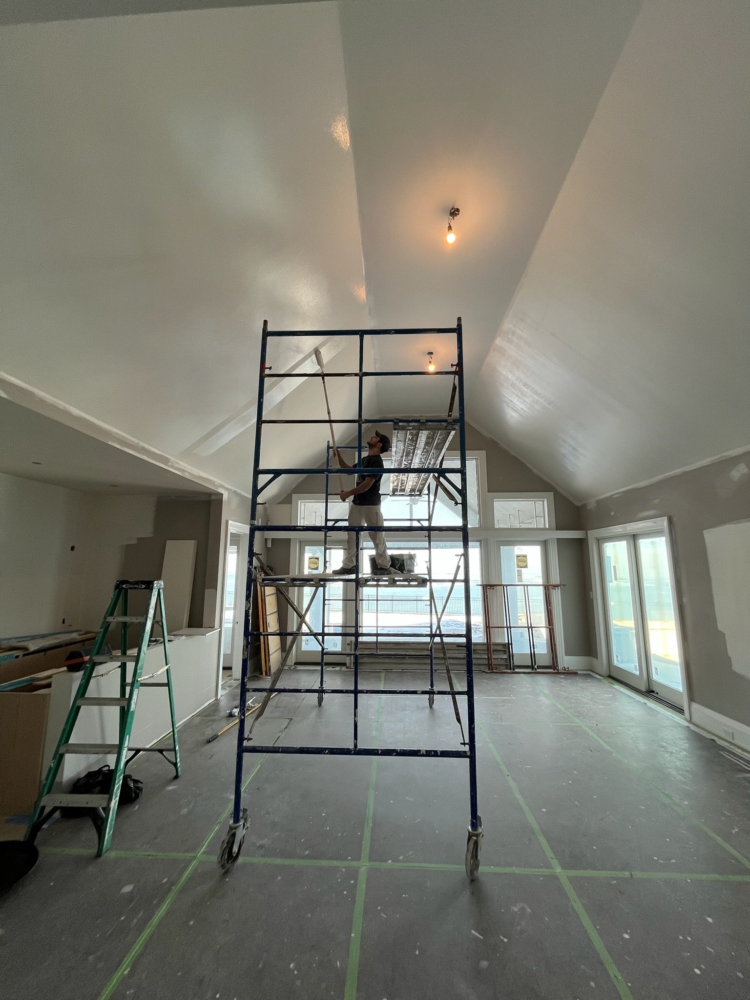 A man painting the ceiling of a large, open room with a roller brush while standing on scaffolding. The room is under construction or renovation, with plastic covering on the window sills and green tape marking the floor. There are additional ladders and construction materials around.