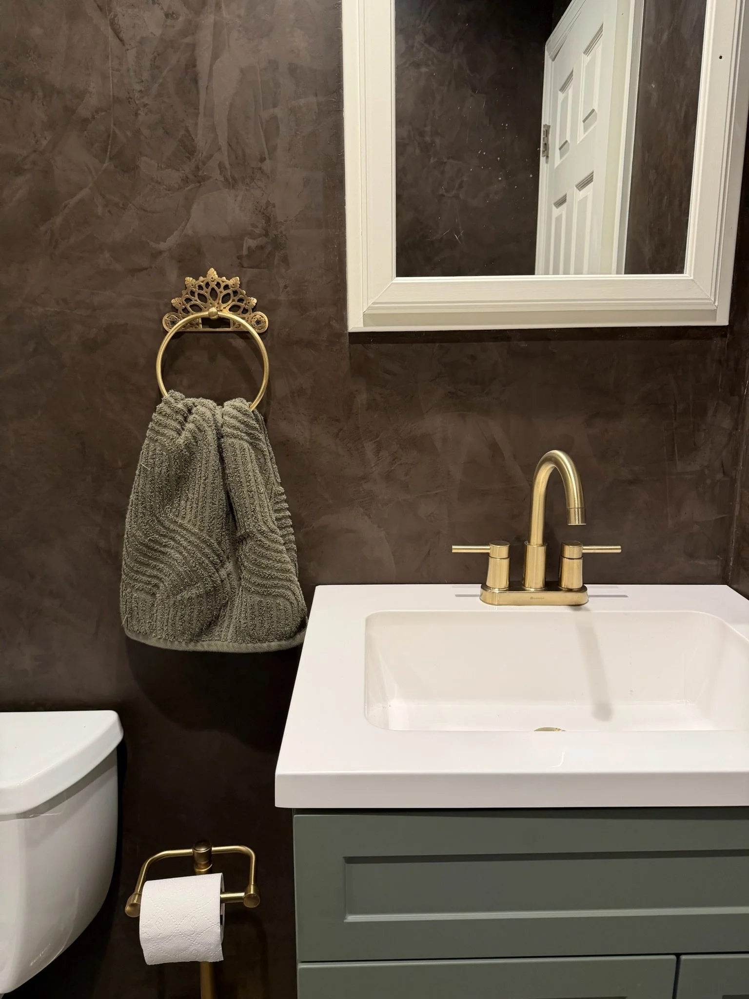 A small bathroom with a dark brown textured wall, gold towel ring with a gray towel, white framed mirror, white sink with gold faucet, and a decorative gold toilet paper holder. Part of a white toilet is visible.