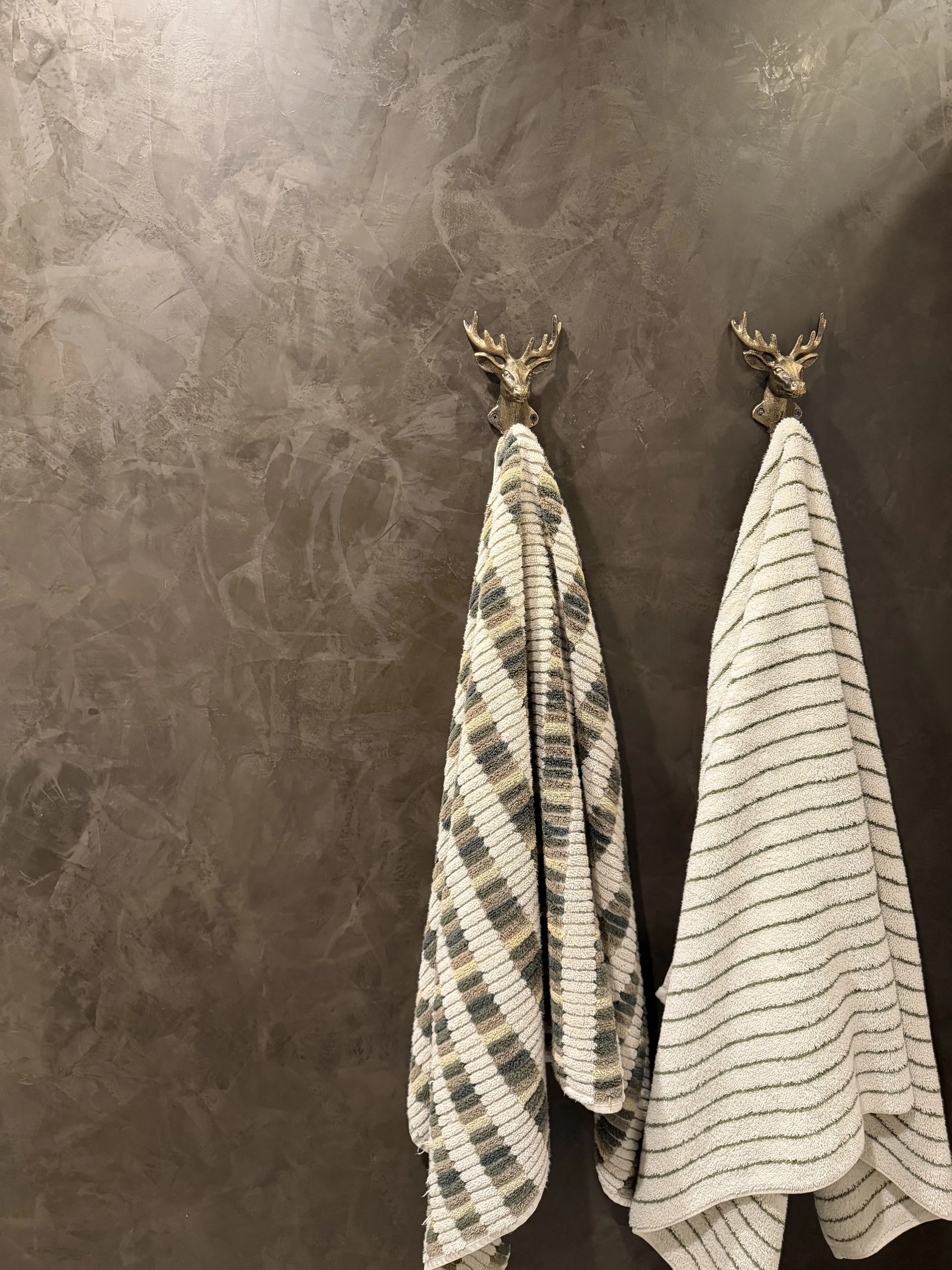 Two towels hanging on wall hooks shaped like deer heads with antlers, mounted on a textured brown wall.