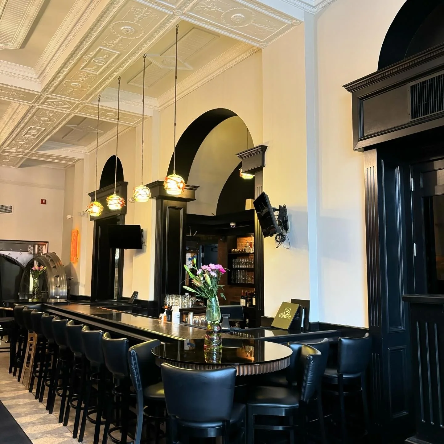 Interior view of a restaurant bar area with black chairs and tables, a vase with pink flowers on the counter, hanging pendant lights, arched doorways, and decorative ceiling details.