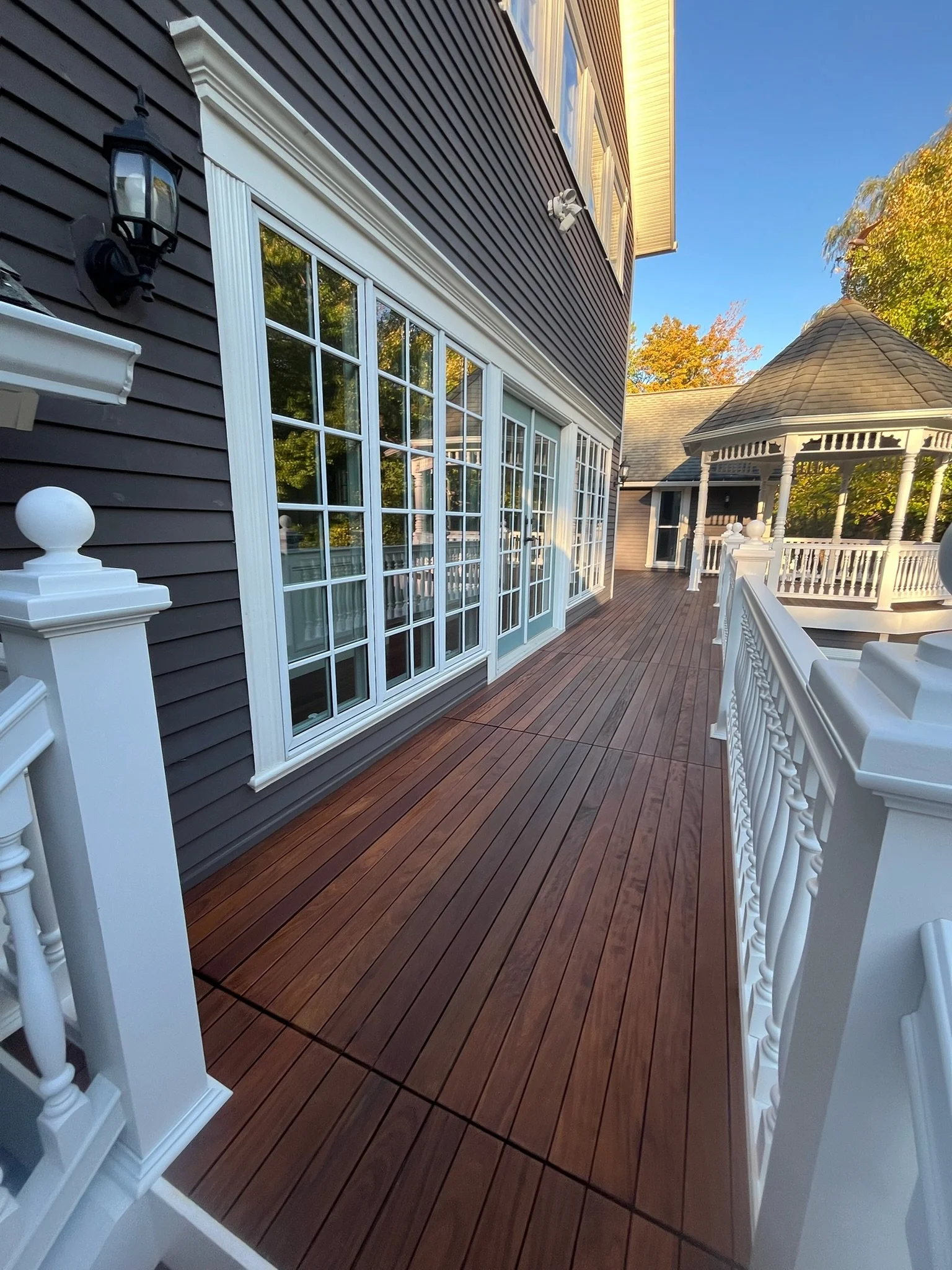 A rear view of a house with a wooden deck, white railing, large glass sliding doors, and a decorative gazebo in the background, with trees and blue sky.