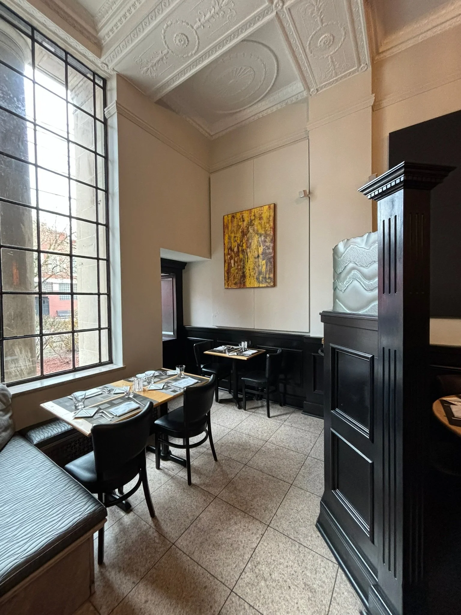Interior of a cozy restaurant corner with a large window, black chairs, set tables with cutlery, glasses, and napkins, a yellow abstract painting on the wall, and ornate ceiling detailing.