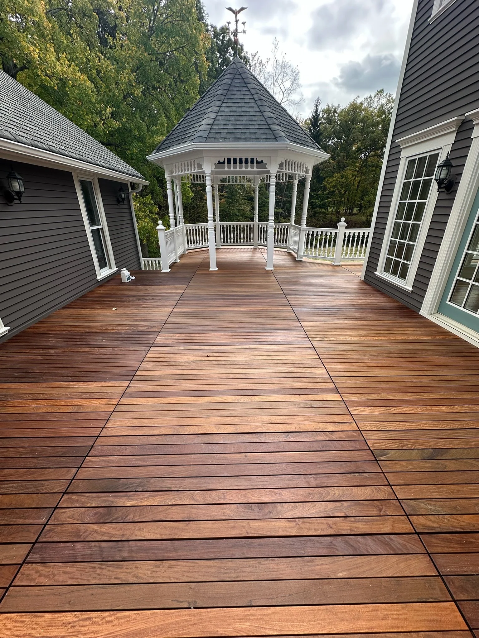 Wooden deck with a white gazebo at the end, surrounded by trees and gray houses with white trim, on a cloudy day.