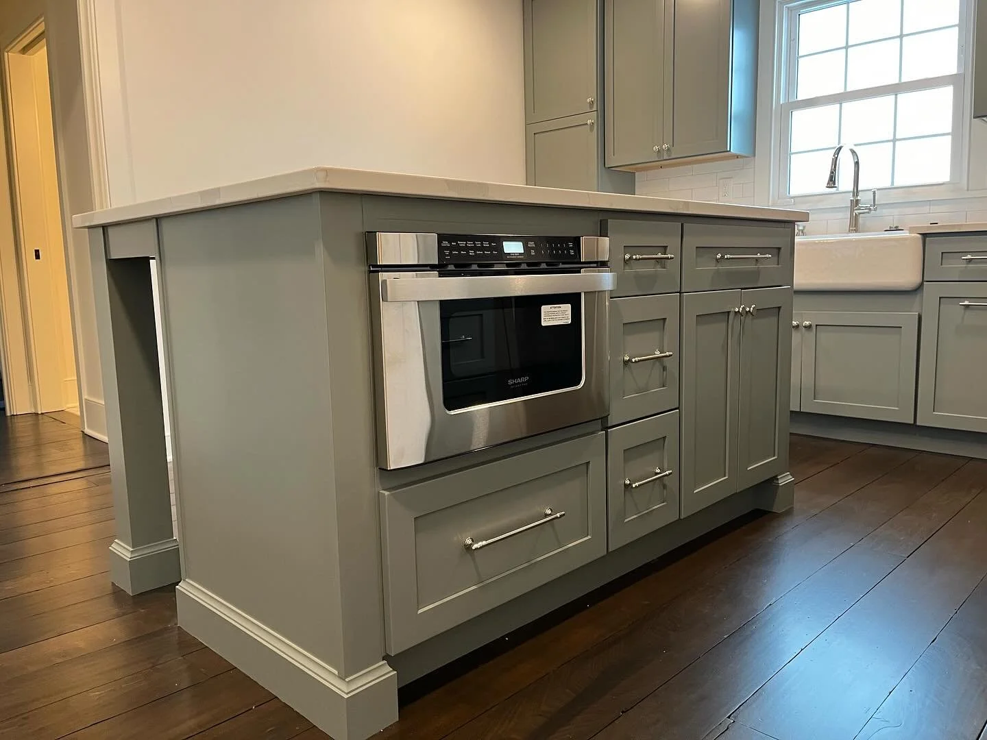Kitchen island with a built-in oven in a modern kitchen with gray cabinets, hardwood floors, and a large window with a farmhouse sink underneath.