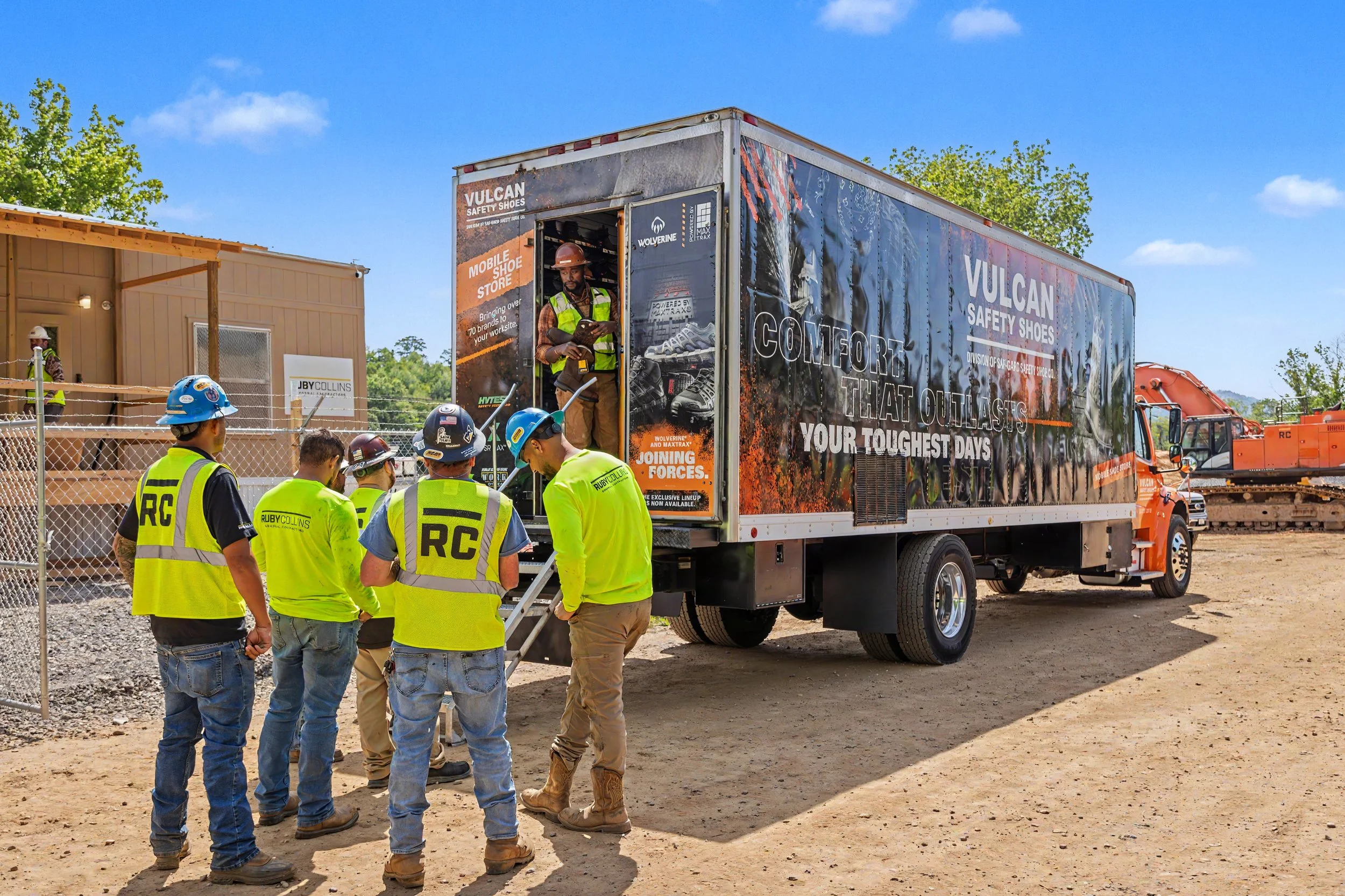 A group of construction workers gathered near a mobile shoe store truck at a construction site on a sunny day. The truck has advertisements for Vulcan safety shoes and features a person inside the truck wearing a safety hat. The workers are wearing yellow safety vests and helmets, and there are construction materials and equipment in the background.