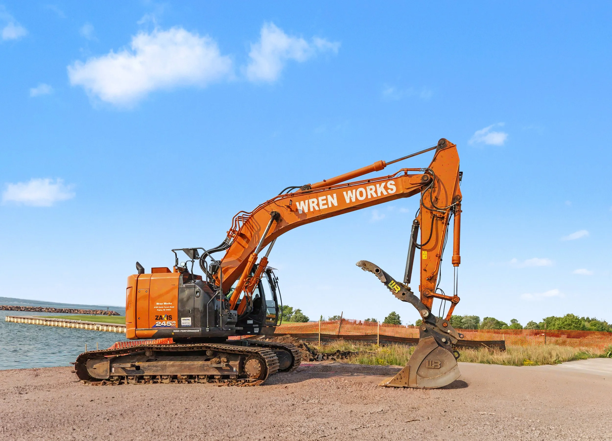 An orange excavator by a body of water under a blue sky with scattered clouds. The excavator has 'WREN WORKS' written on its arm and is on a gravel surface, possibly at a construction site.