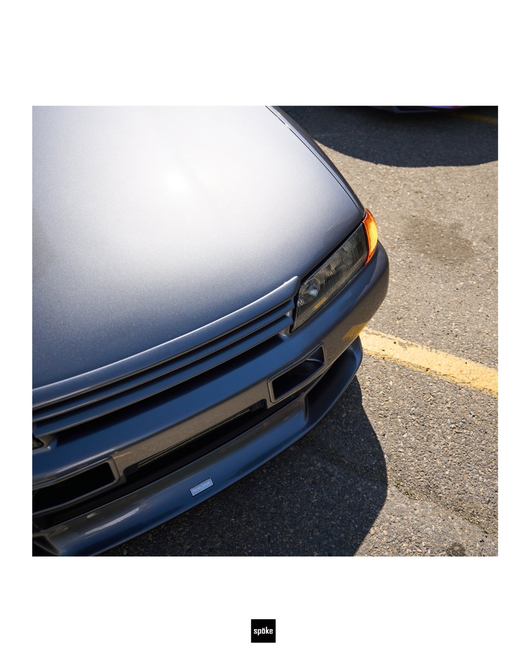 Close-up of the front end of a silver sports car parked on an asphalt surface, showing part of the hood, headlight, and front bumper.