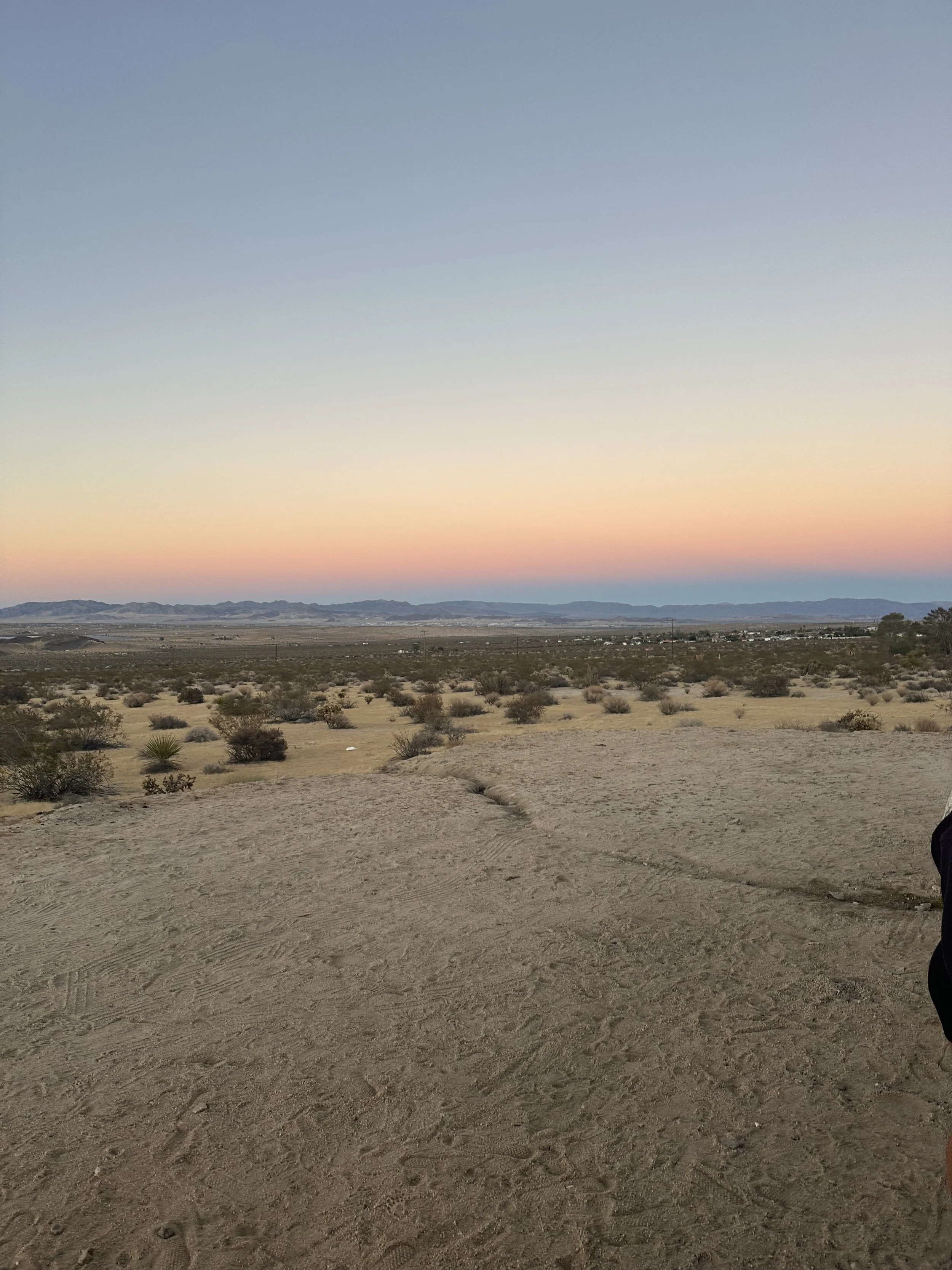 Desert landscape at sunset with dry dirt ground, sparse bushes, mountains in the distance, and a colorful sky with pastel hues.