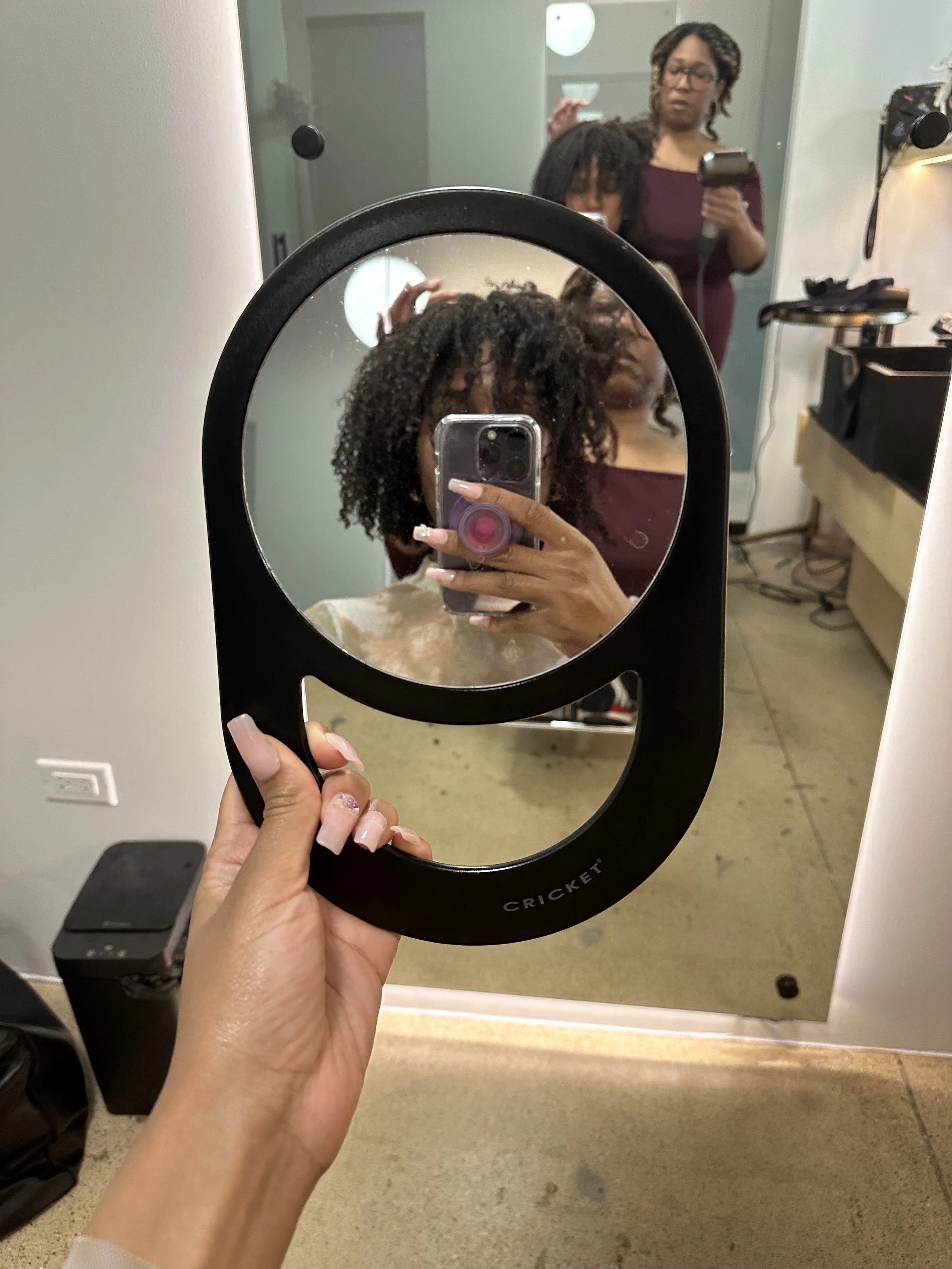 a black woman with curly hair holding a mirror up while getting her hair done